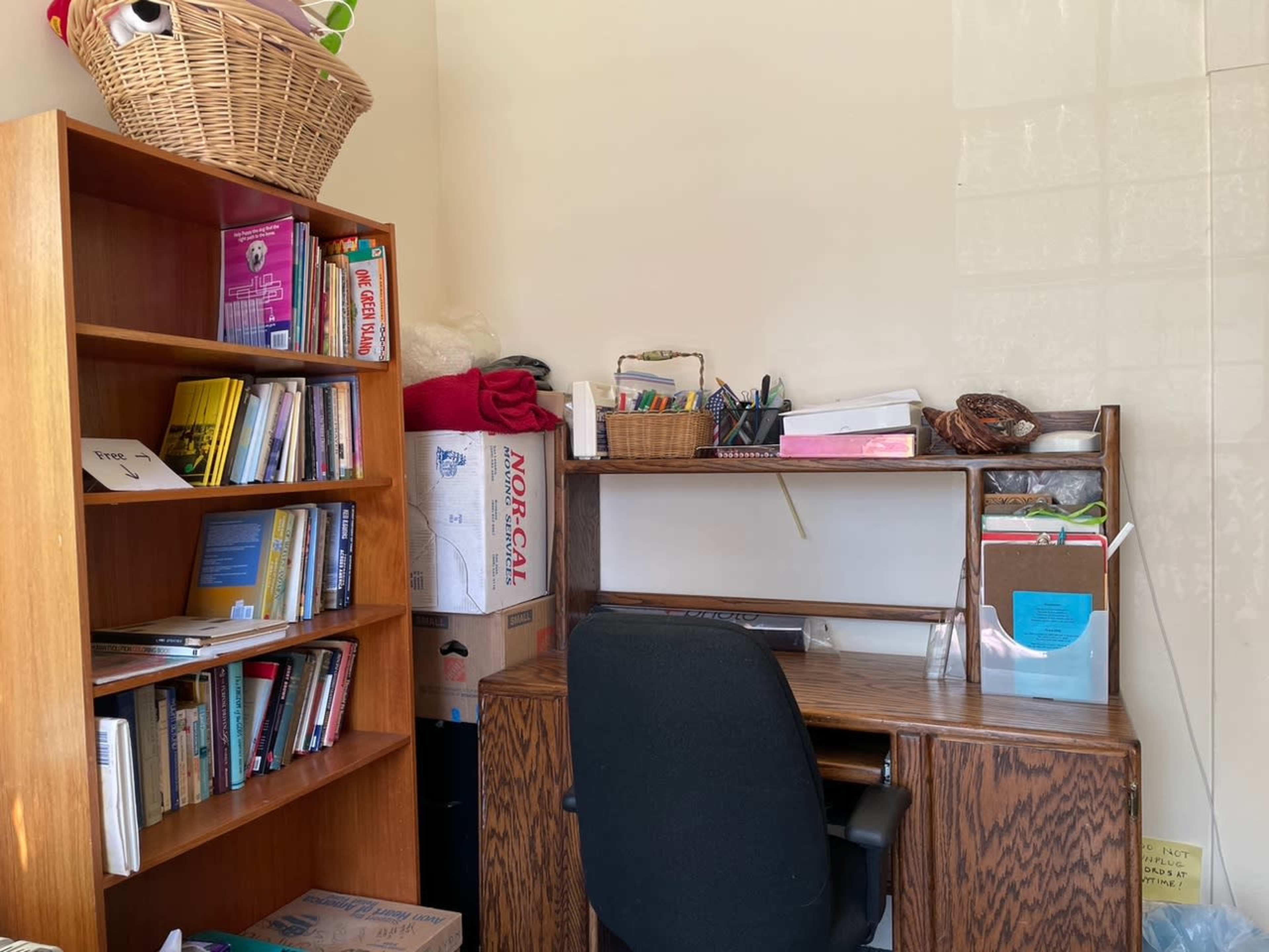 The image shows a small home office area with a wooden desk and chair beside a bookshelf filled with books and storage boxes.