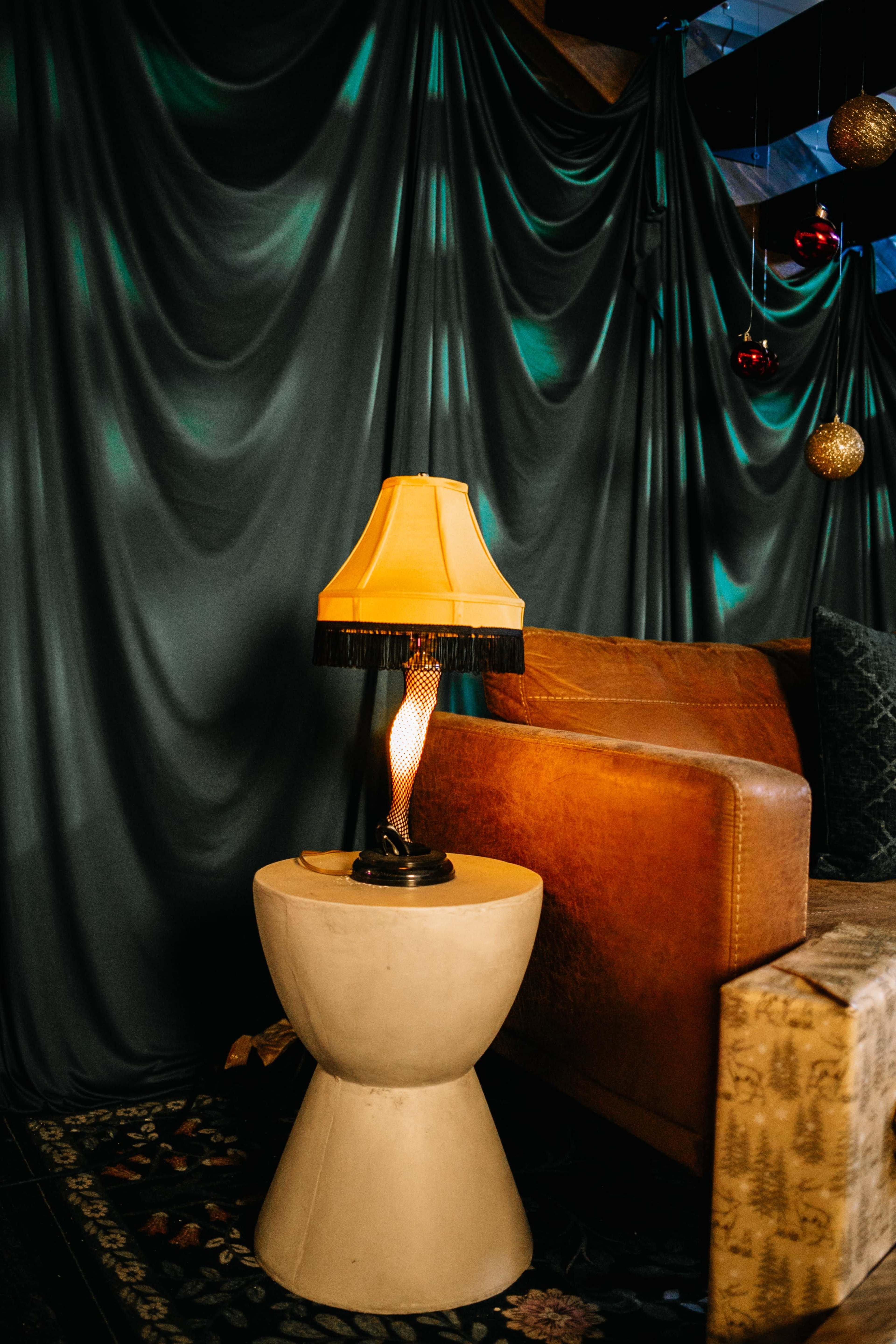 A modern lamp sits on a round, light-colored side table next to a leather sofa, with a draped fabric backdrop featuring decorative ornaments.