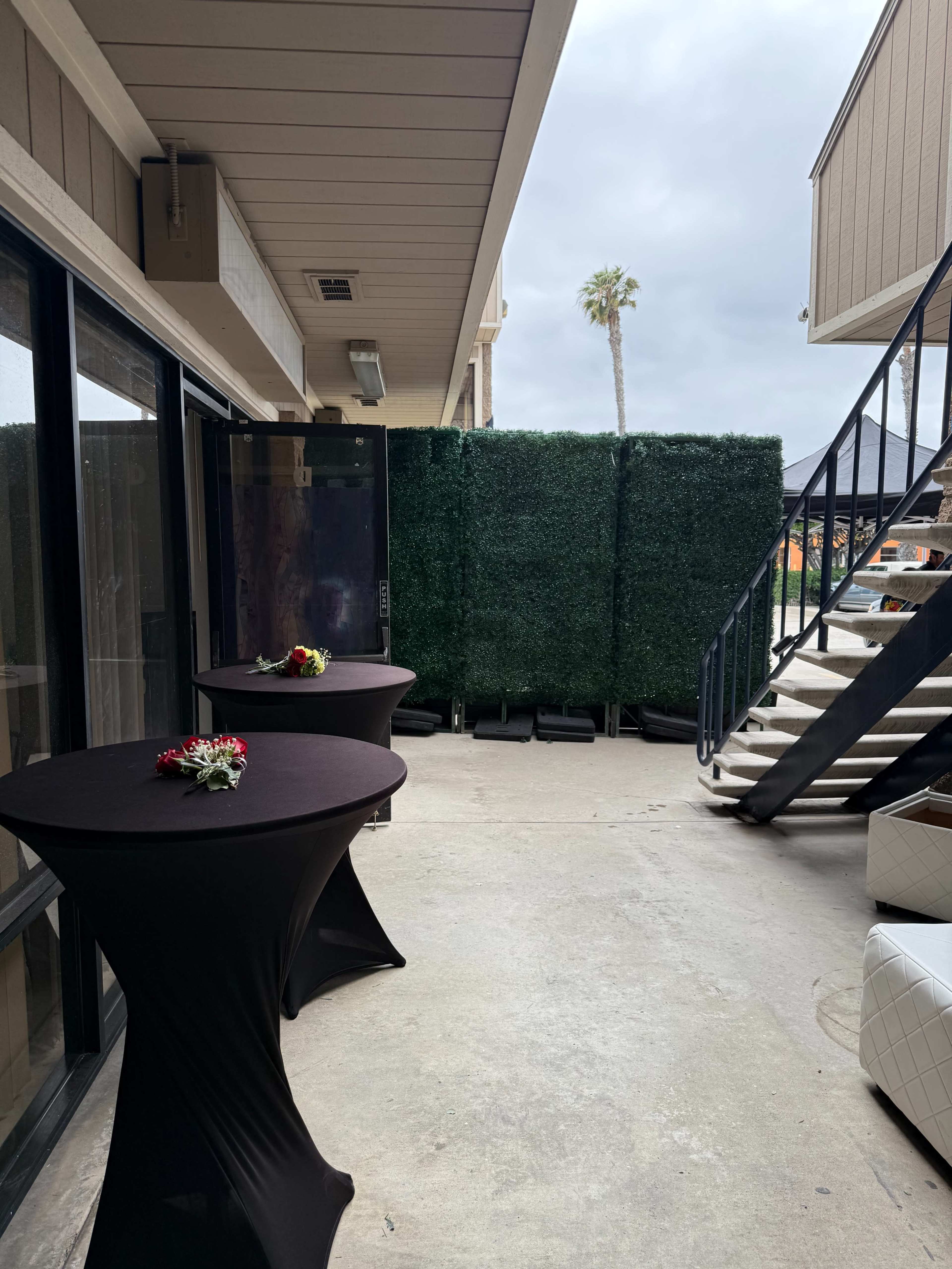 The image shows a courtyard area with two black tablecloth-covered tables, decorated with flowers, and a backdrop of greenery near a staircase and a building exterior.