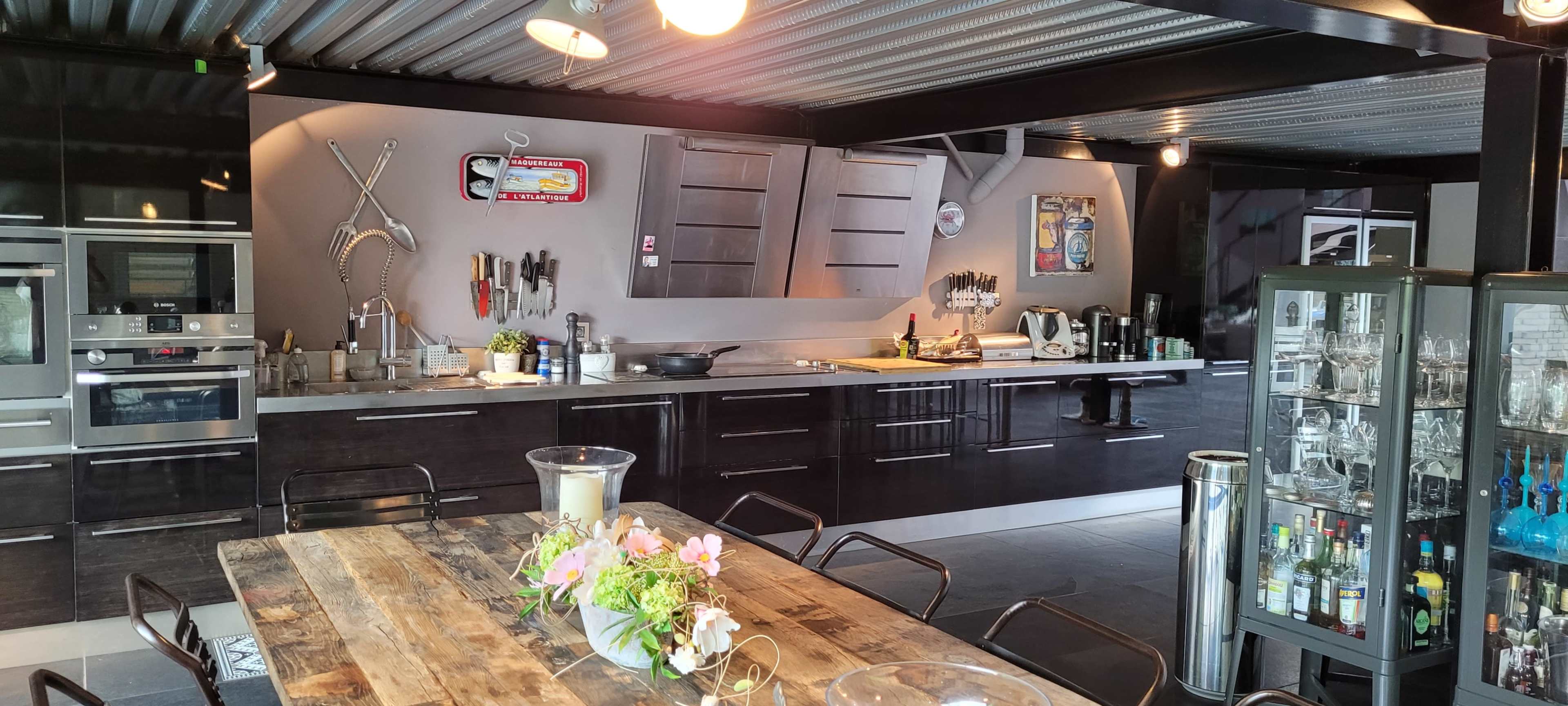 A modern kitchen with a sleek black cabinetry, a long wooden dining table with flowers, and various kitchen tools displayed on the wall.