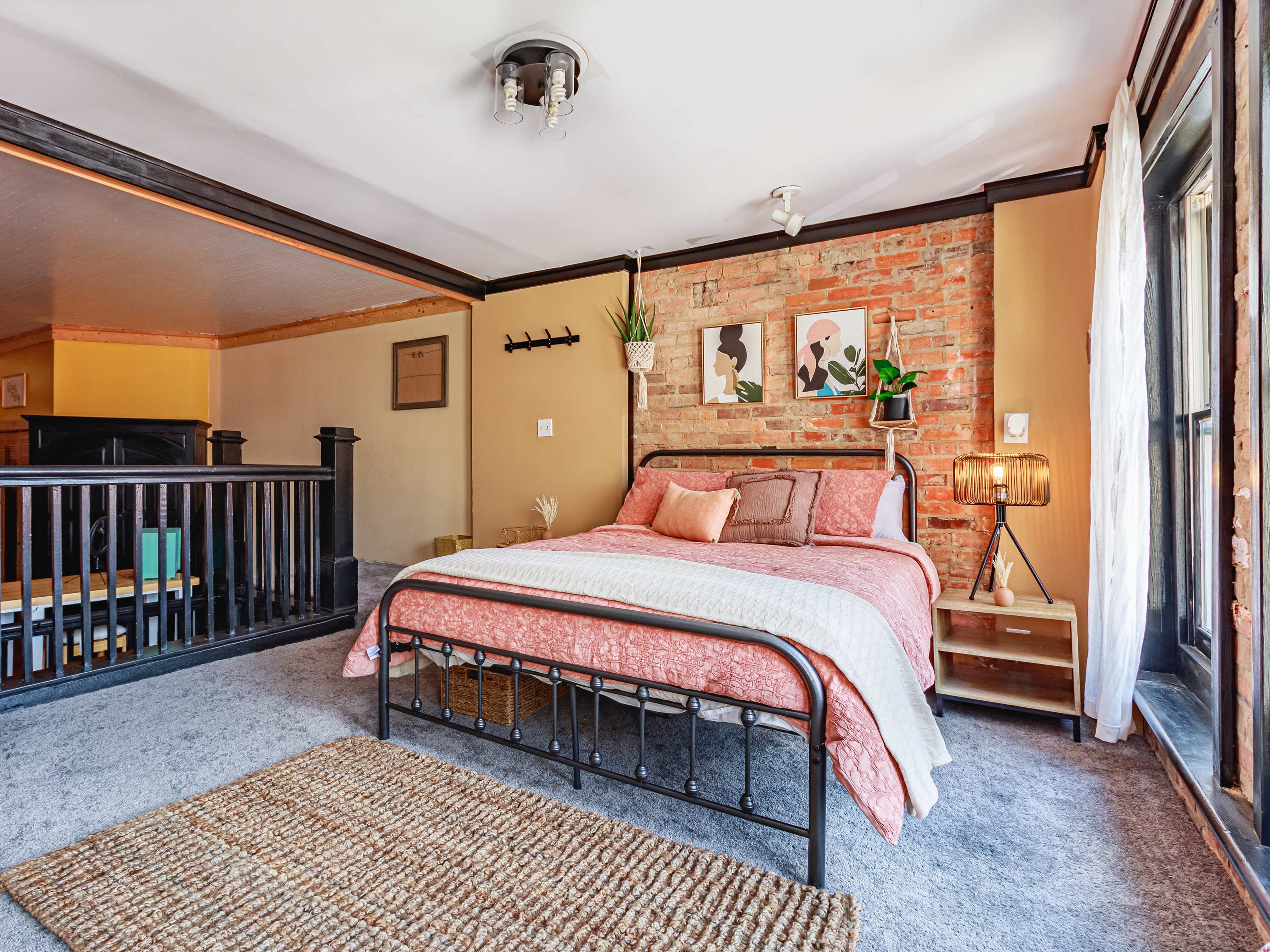 The image shows a bedroom featuring a metal bed frame with a patterned bedspread, a brick accent wall, and a wooden railing.