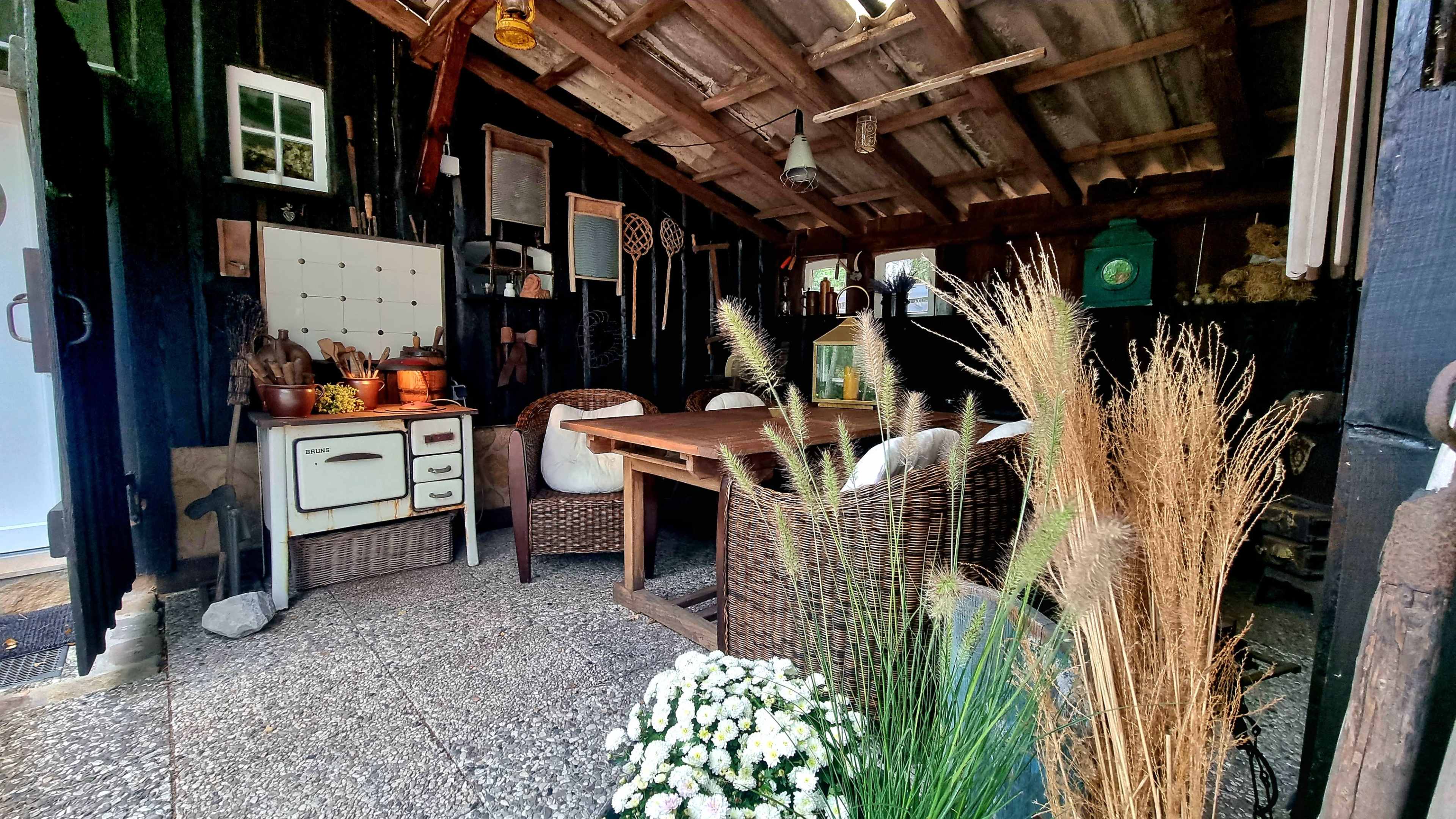 The image shows a rustic shed interior with a wooden table, wicker chairs, and vintage kitchen items arranged along the walls.