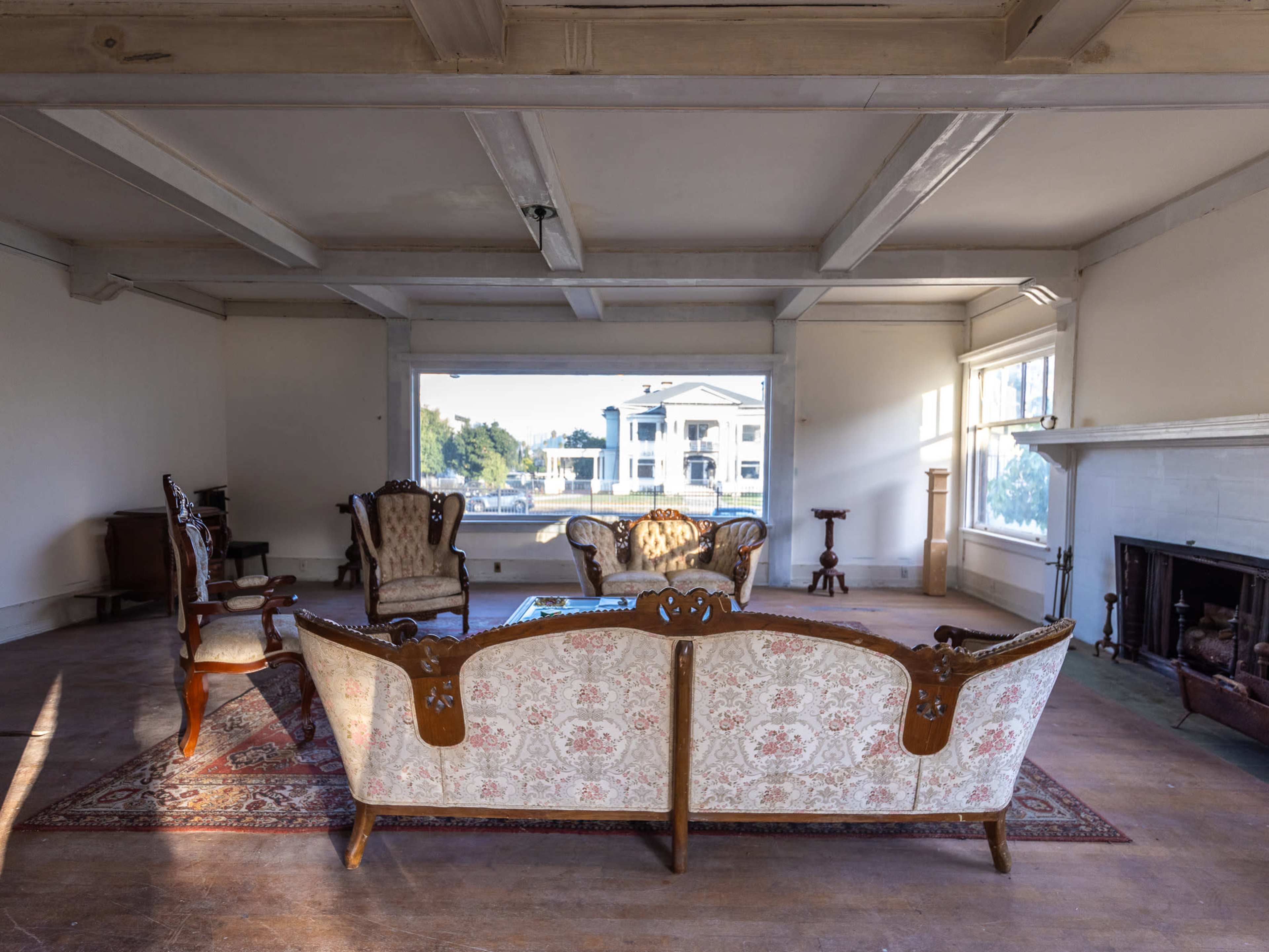A spacious, empty living room features vintage furniture, including a central floral-patterned sofa, two armchairs, and a wooden table, with large windows illuminating the space.