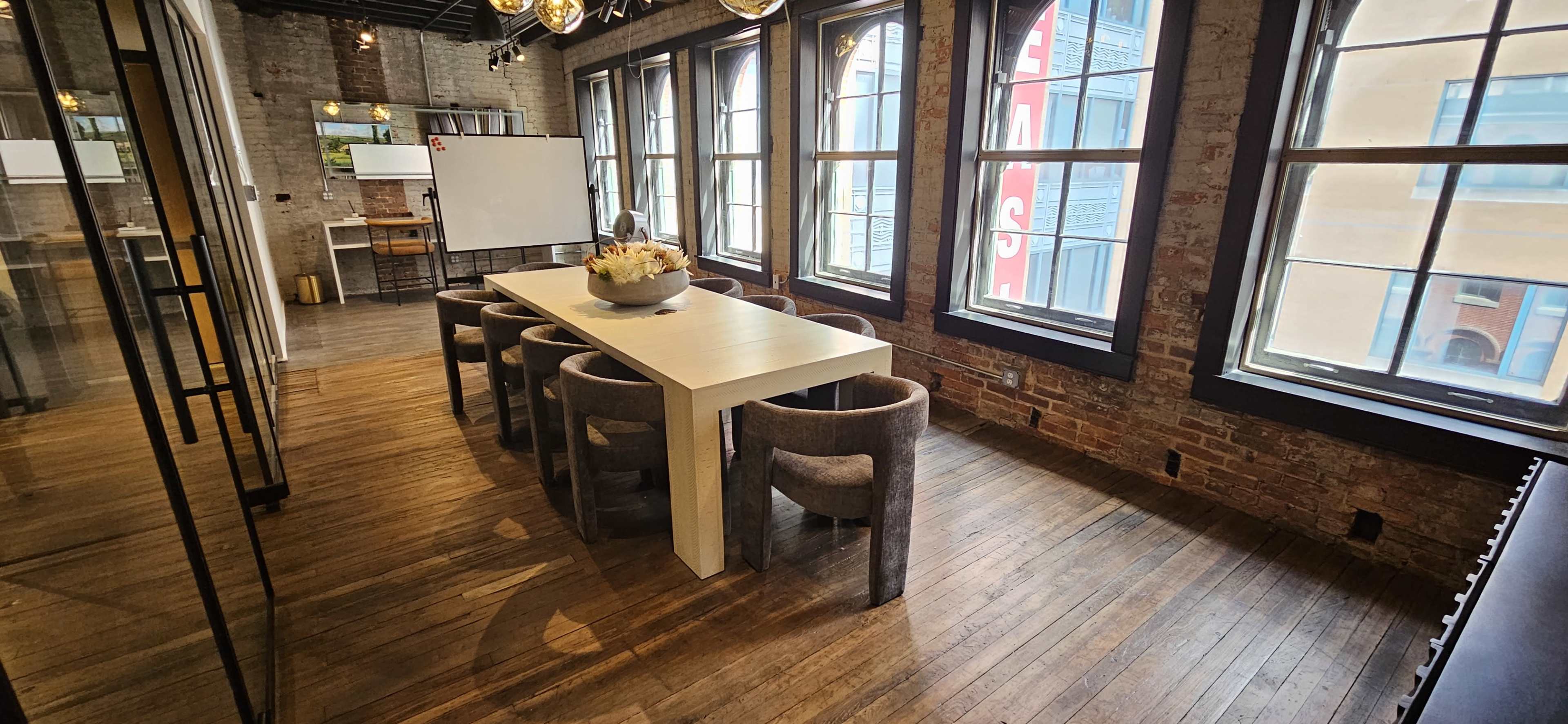 A long table surrounded by chairs is set in a well-lit meeting room with large windows and exposed brick walls.