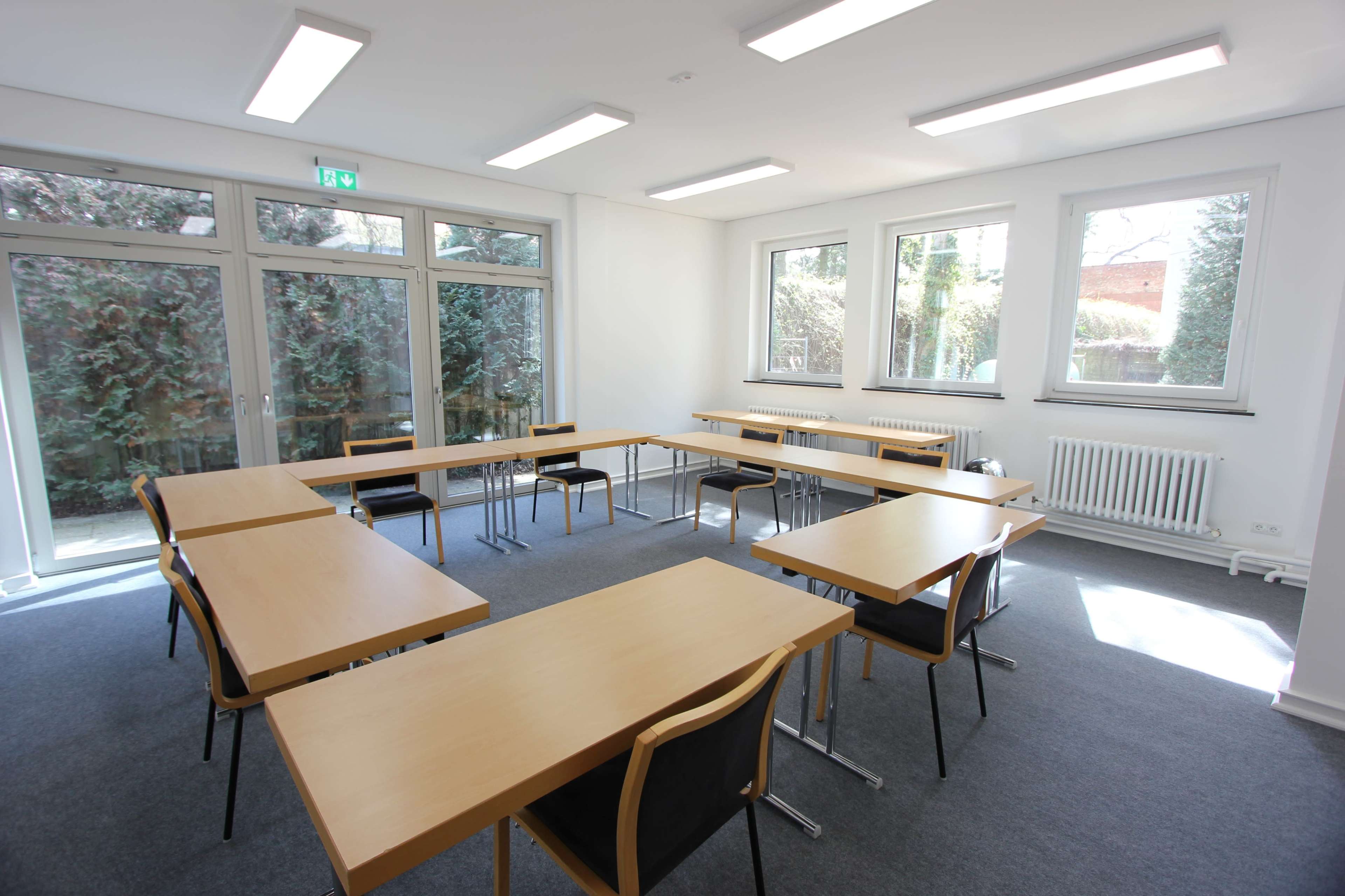 The image shows a bright, empty classroom with multiple tables and chairs arranged in a U-shape, surrounded by large windows.