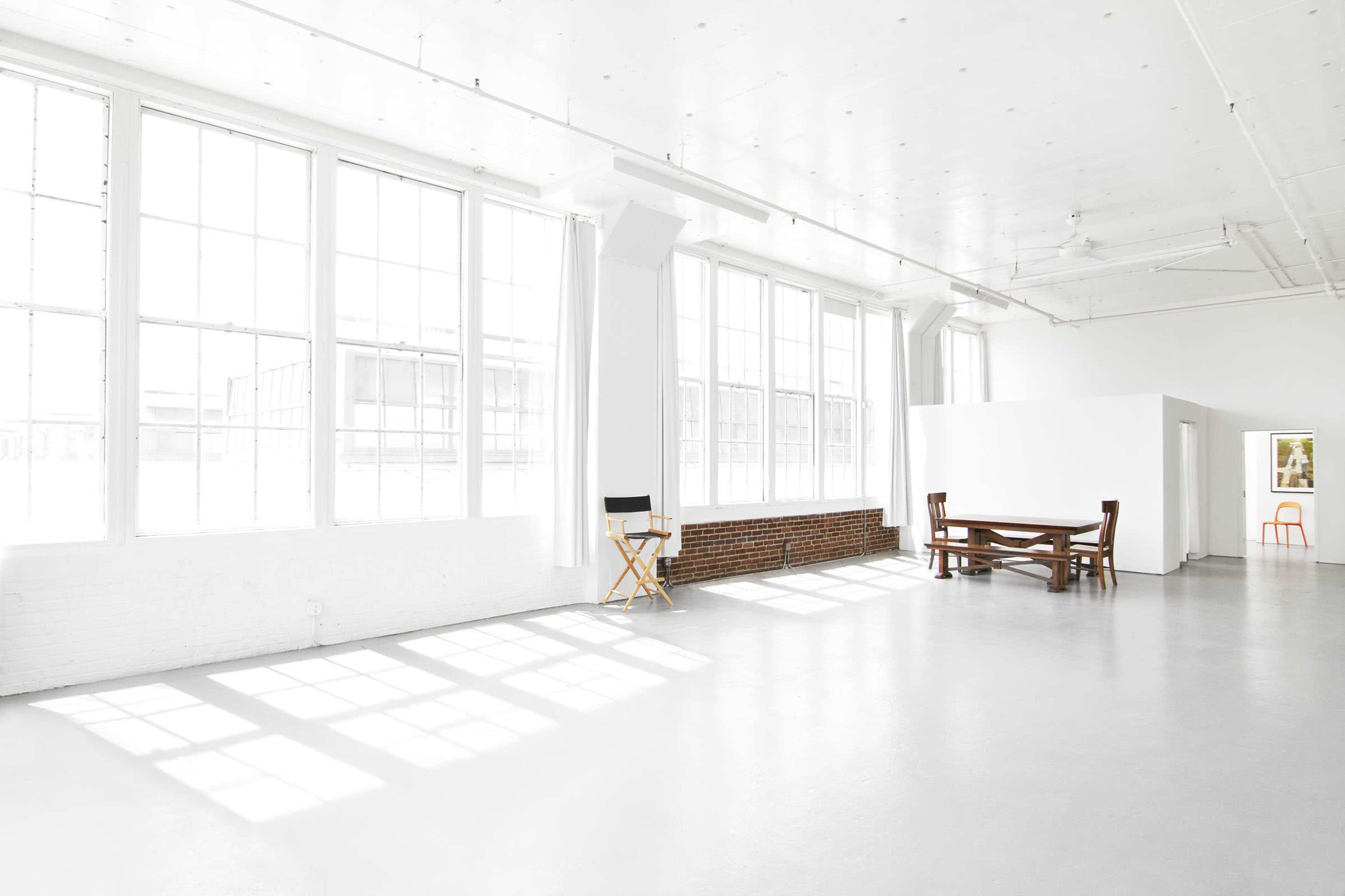 A spacious, bright room with large windows, a lone director's chair, a wooden table, and a white partition against a brick wall.