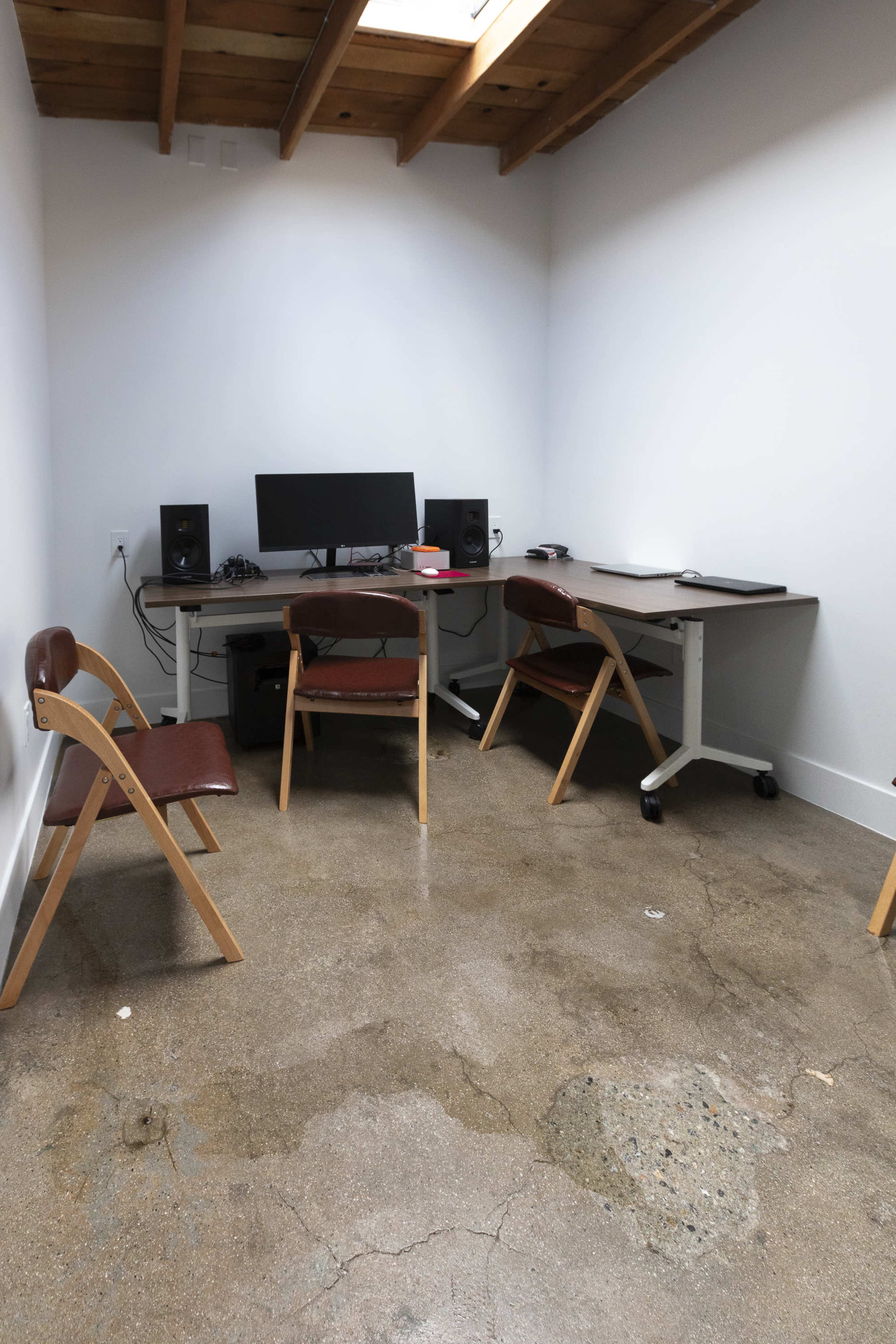 The image shows an empty workspace with a desk, computer monitor, speakers, and several wooden chairs in a room with a polished concrete floor.