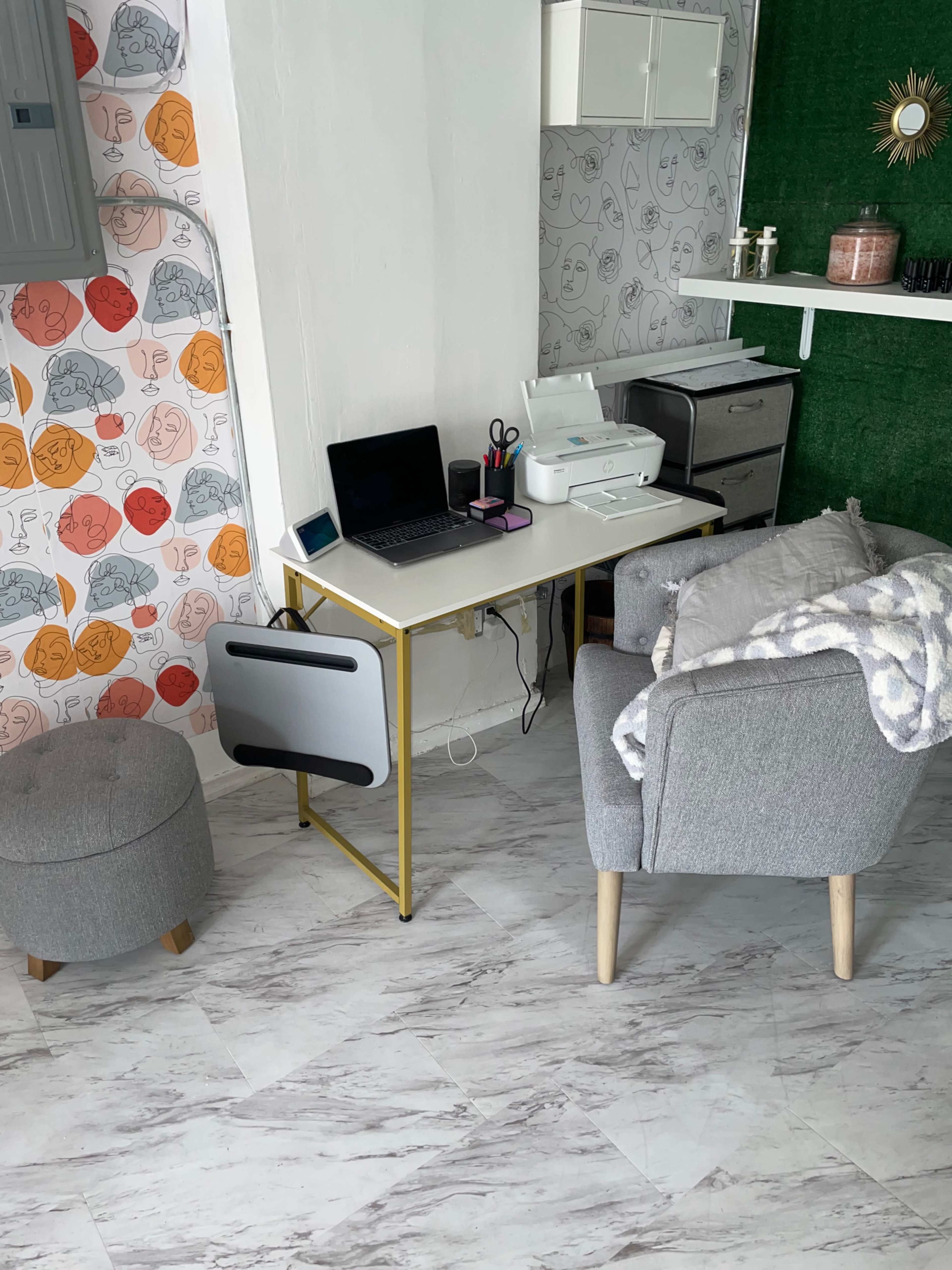 The image shows a small workspace featuring a desk with a laptop, printer, and office supplies, accompanied by a gray armchair and an ottoman, against a patterned wall and a green accent wall.