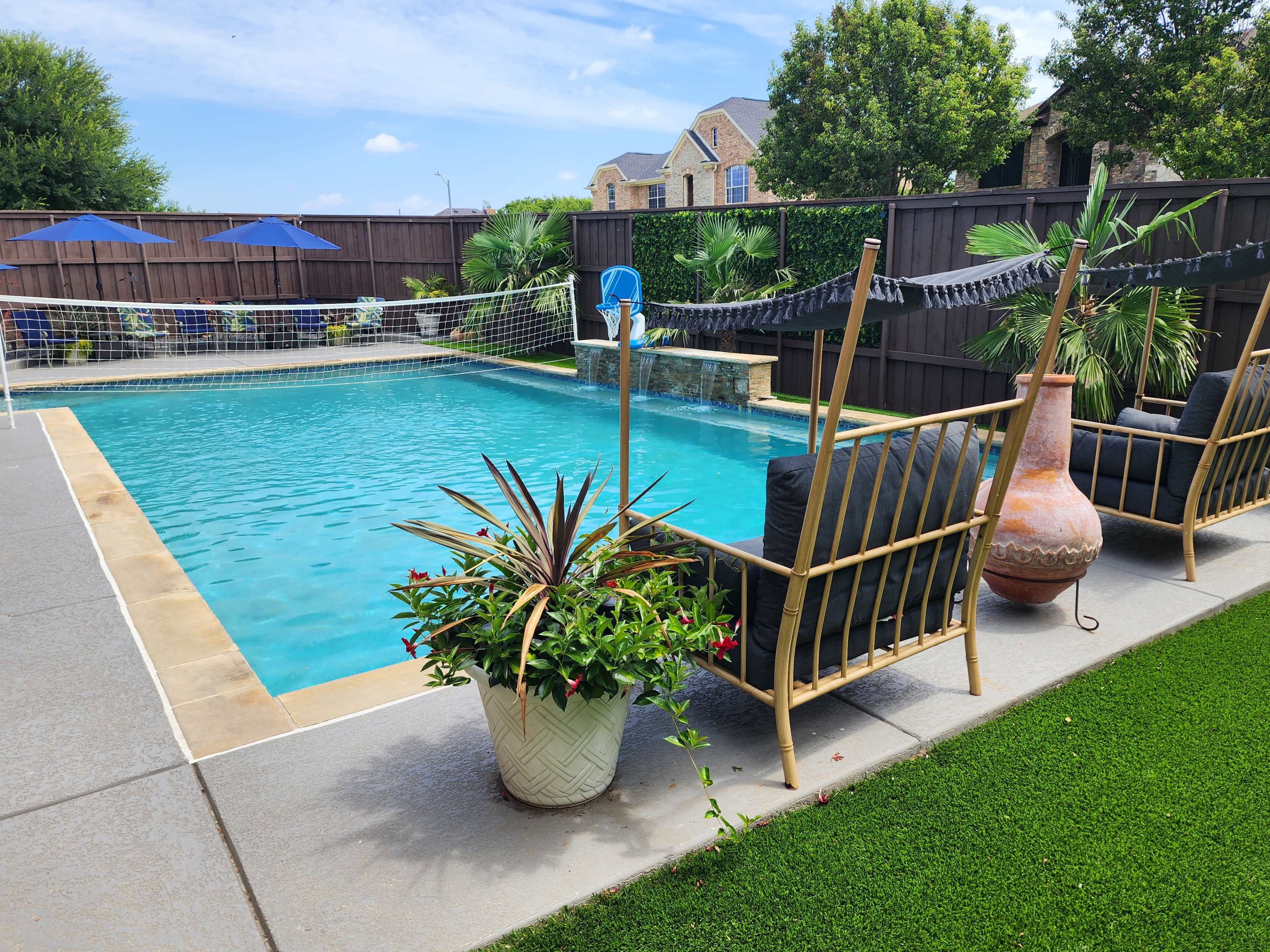 The image shows a swimming pool surrounded by lounge chairs, potted plants, and a volleyball net, with a residential fence and houses in the background.