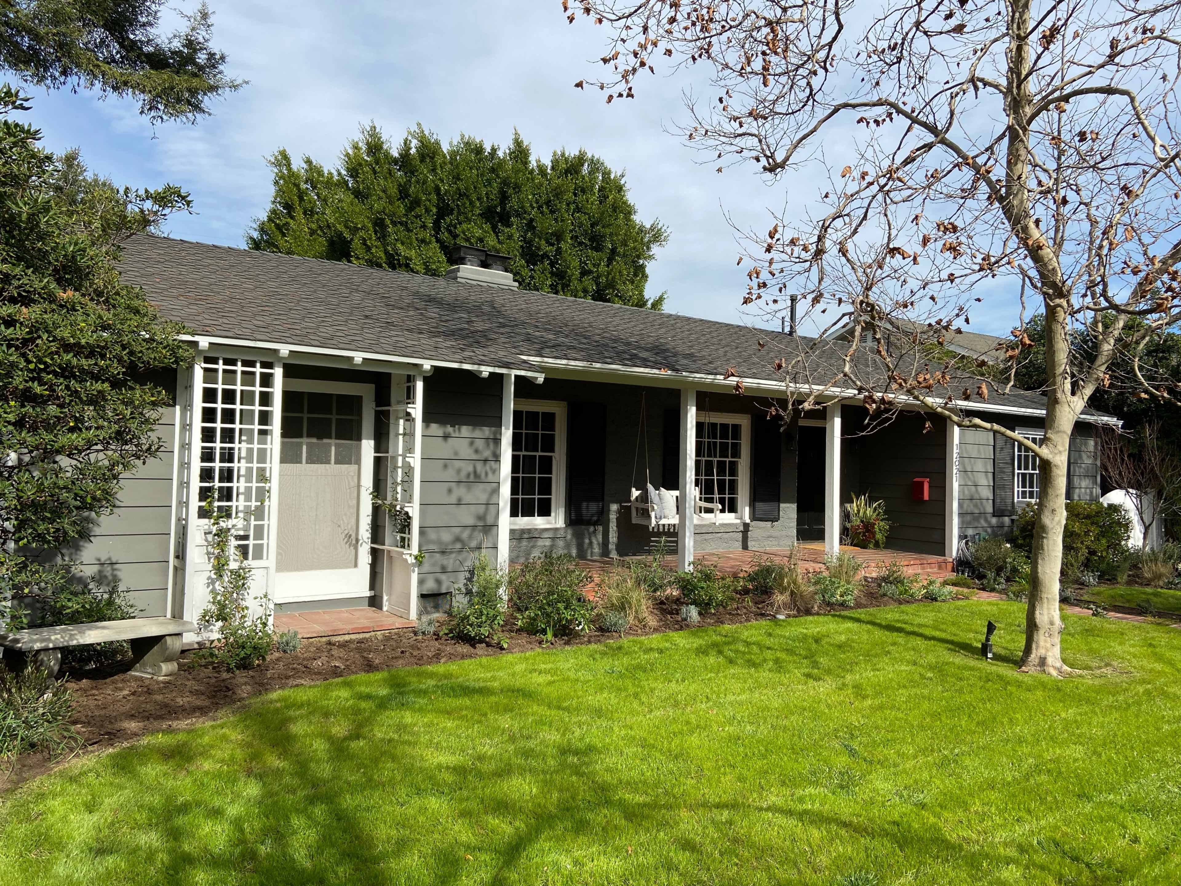A single-story gray house with a brick path and a well-maintained lawn features a front porch and a large tree in the yard.