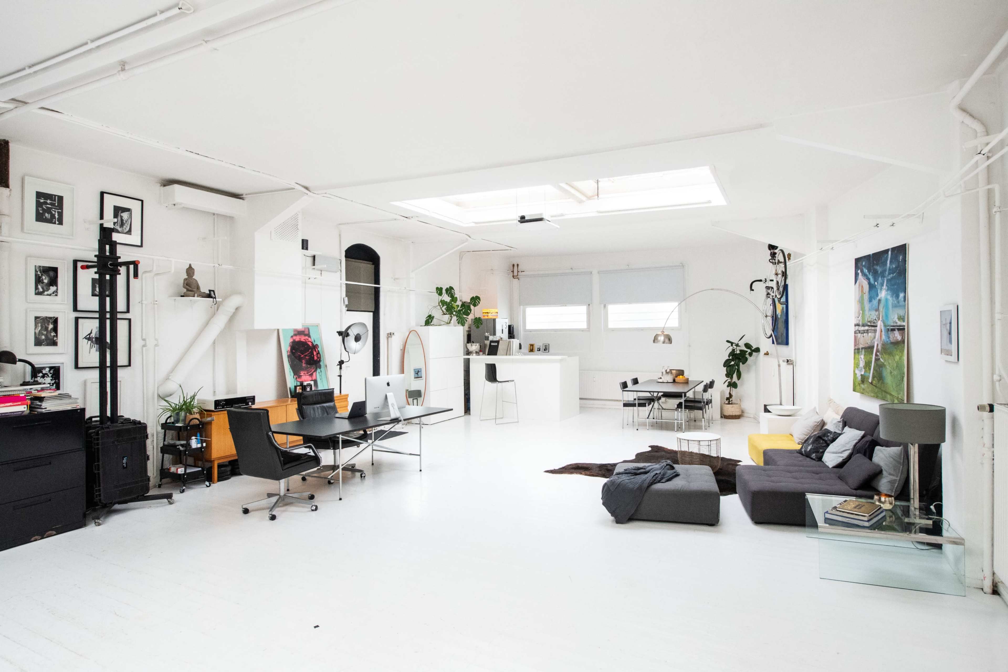 The image shows a spacious, modern loft with white walls, a mix of furniture including a sofa and office chairs, and large windows allowing natural light to enter.