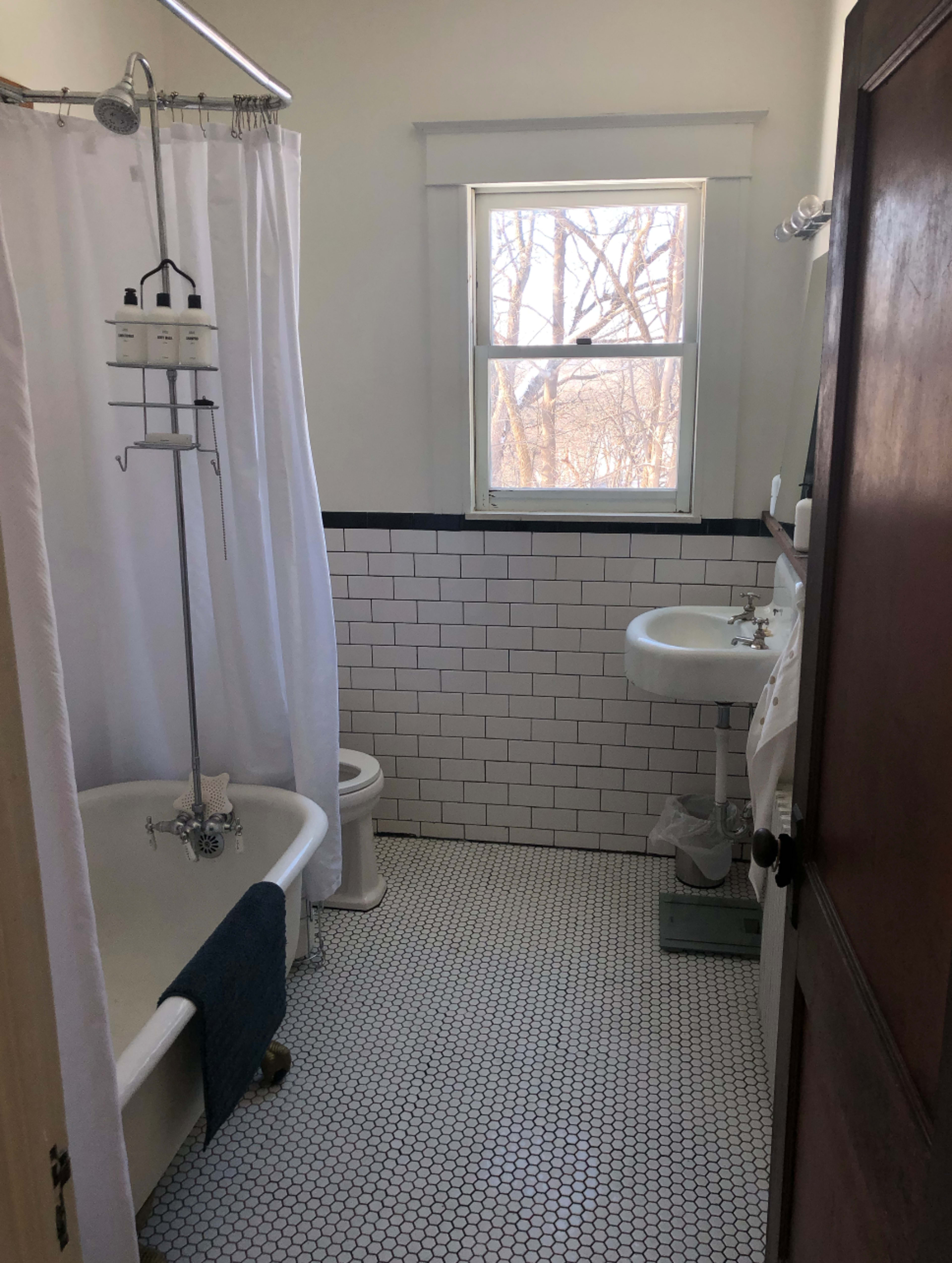 The image shows a bathroom featuring a clawfoot bathtub, a white shower curtain, a sink, a toilet, and a window with a view of bare trees outside.