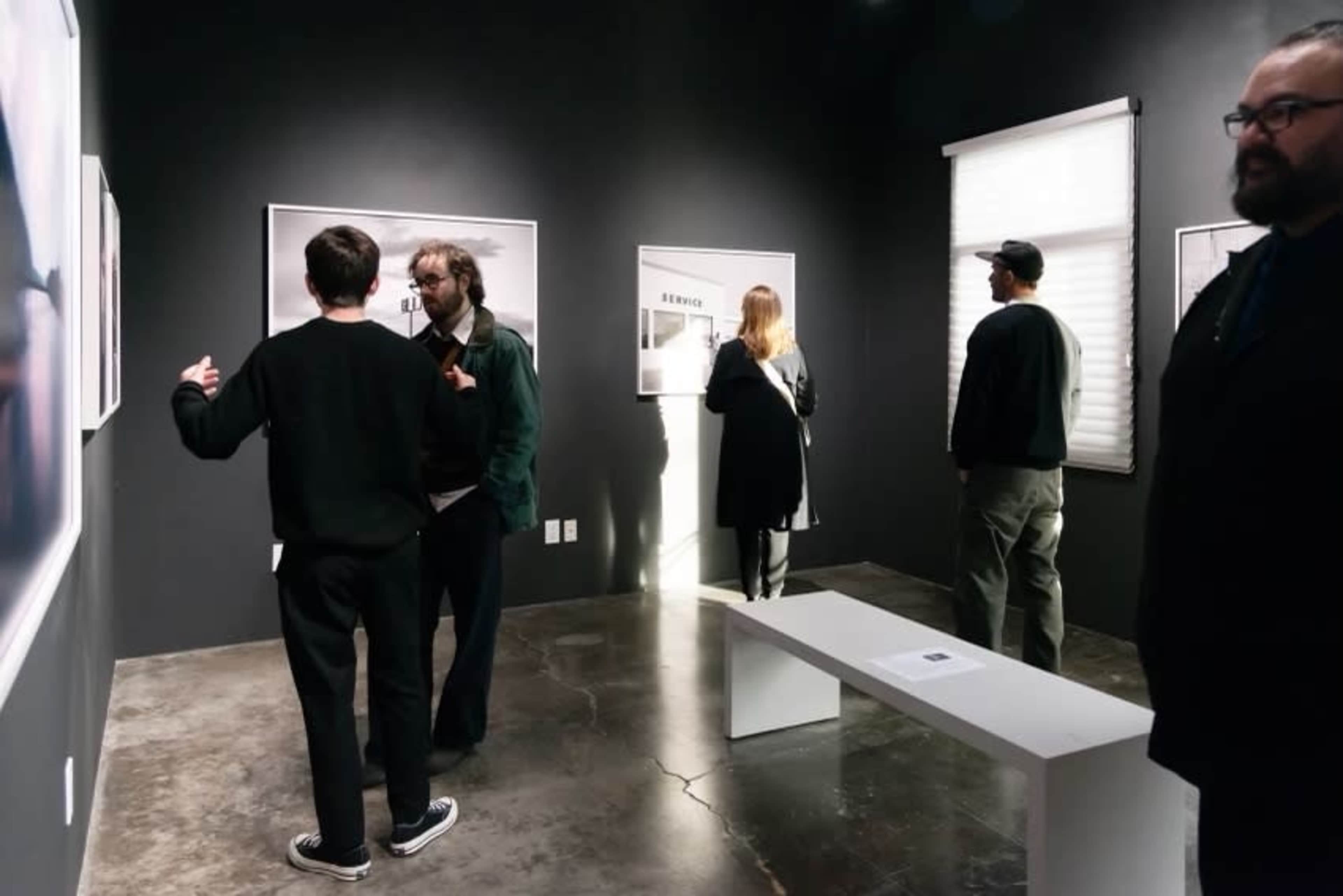 A group of people observes artwork in a dimly lit gallery with black walls and polished concrete floors.