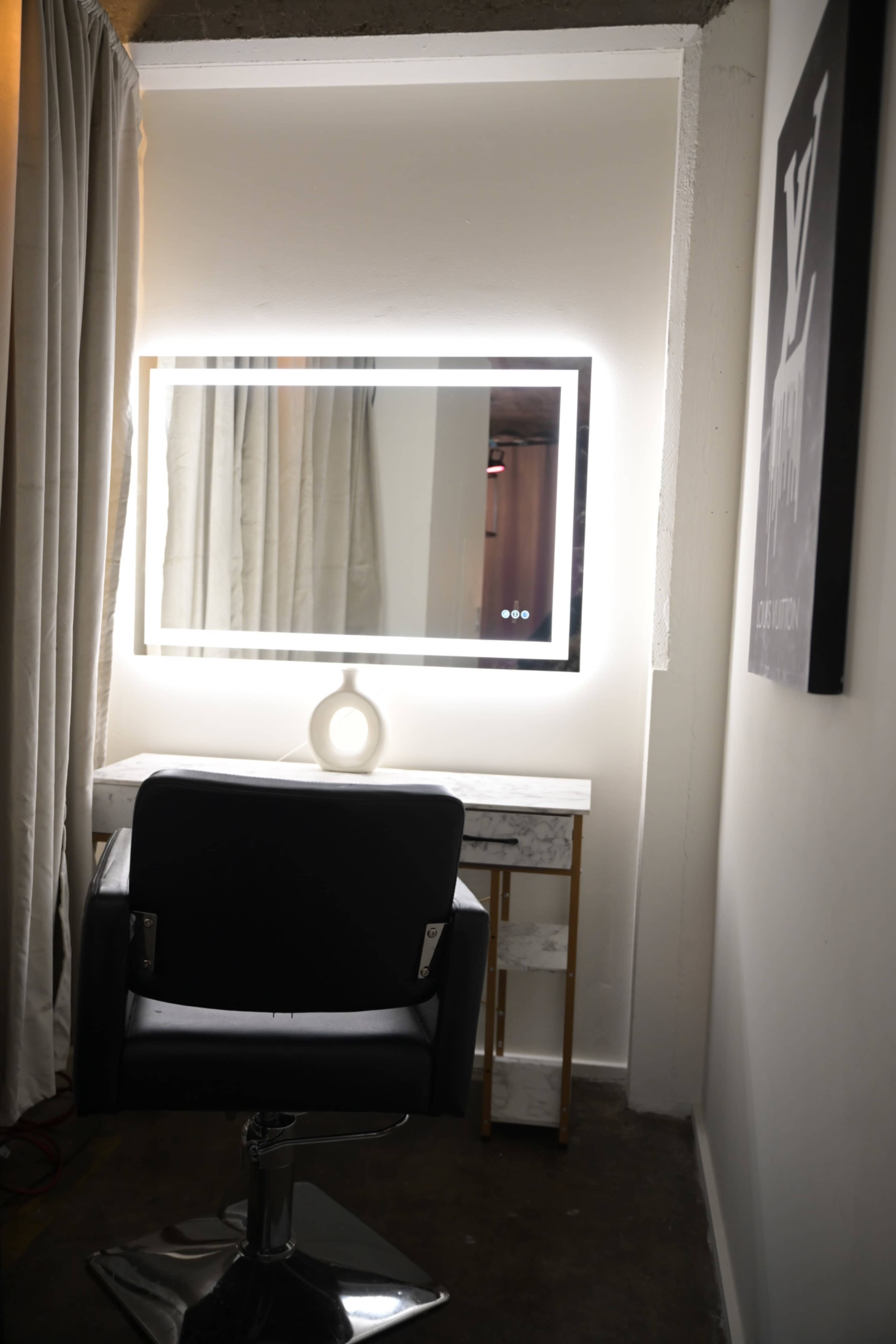 The image shows a well-lit vanity area featuring a large illuminated mirror, a modern chair, and a small table with a lamp against a neutral wall.