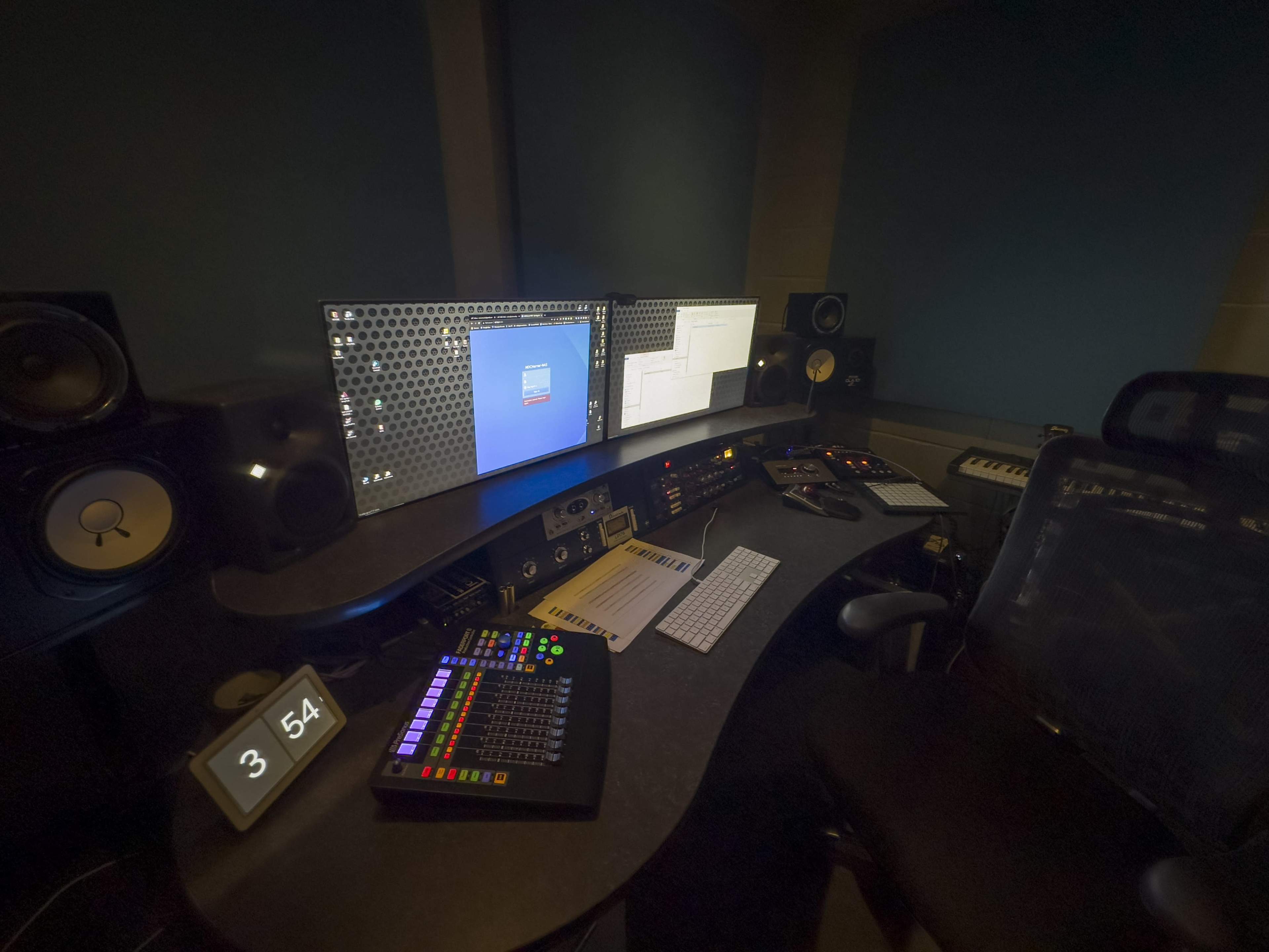 A dimly lit music production studio with multiple computer screens, audio equipment, and a keyboard arranged on a curved desk.