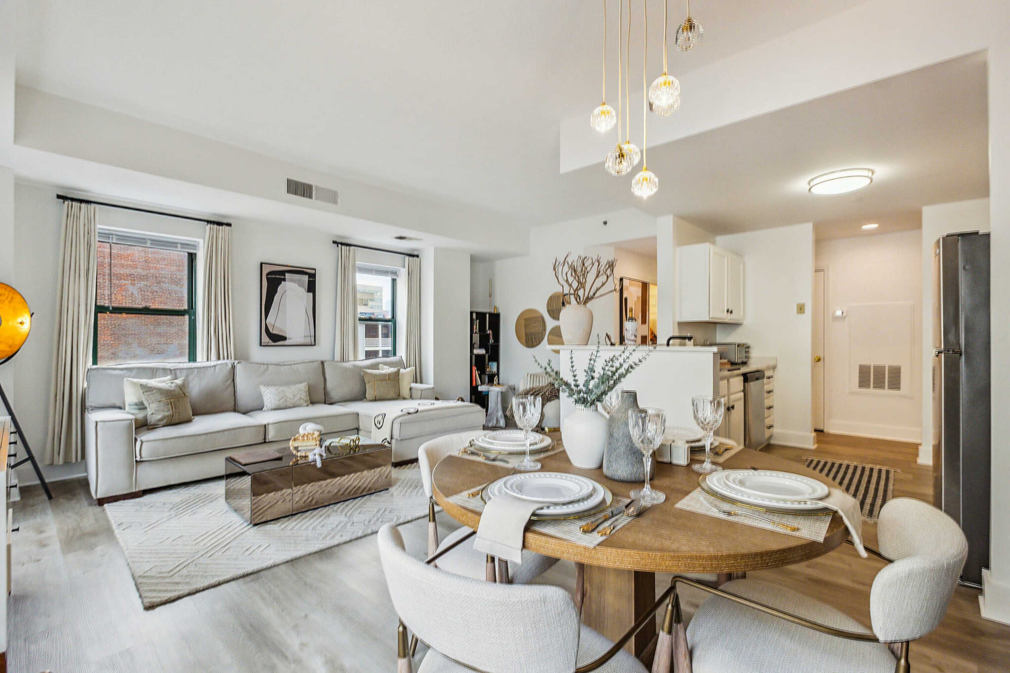 A modern living space featuring a beige sectional sofa, a round dining table set for four, and an open kitchen area with white cabinetry.