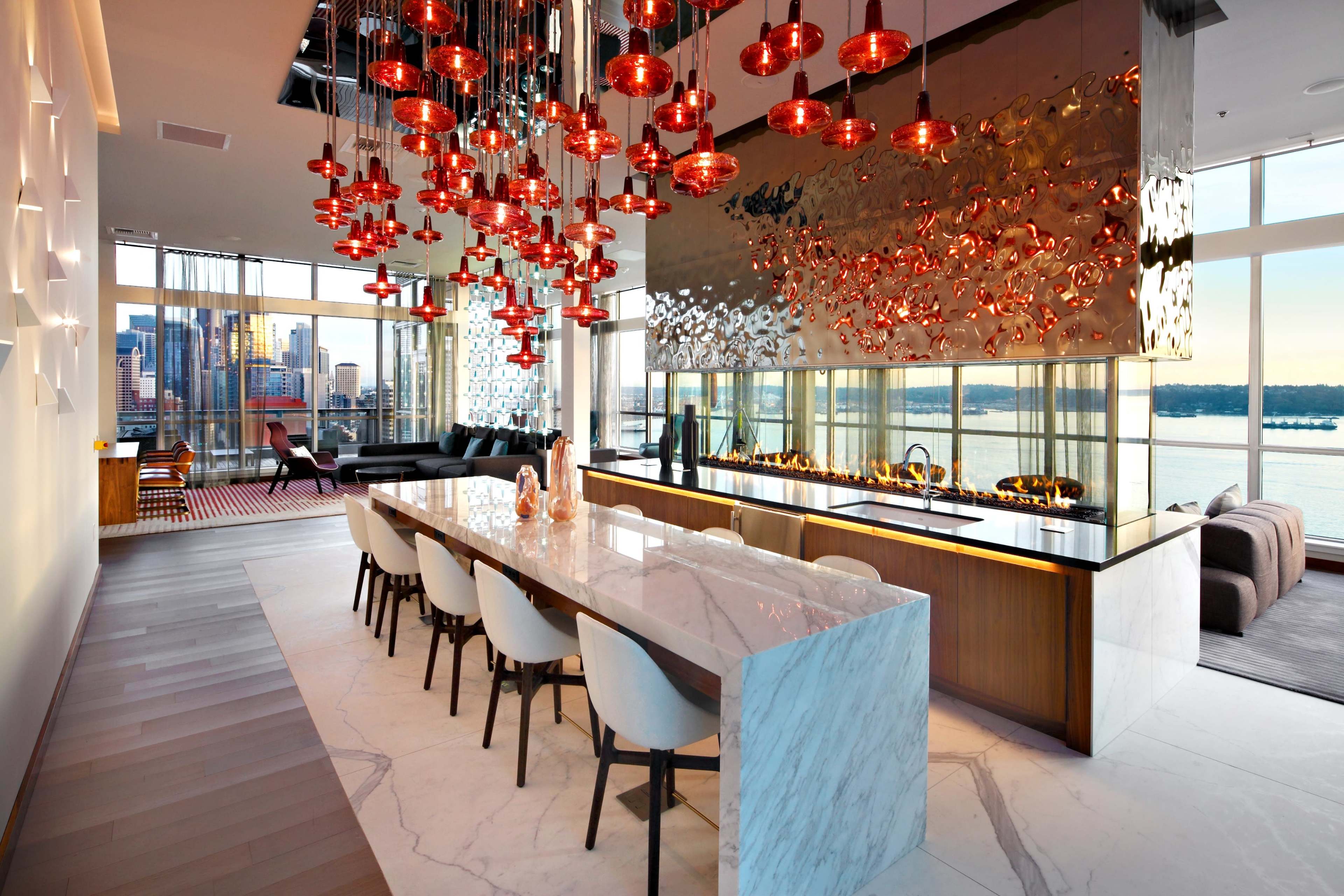 A modern kitchen features a long marble island with seating, surrounded by stylish pendant lights and large windows showcasing a city view.
