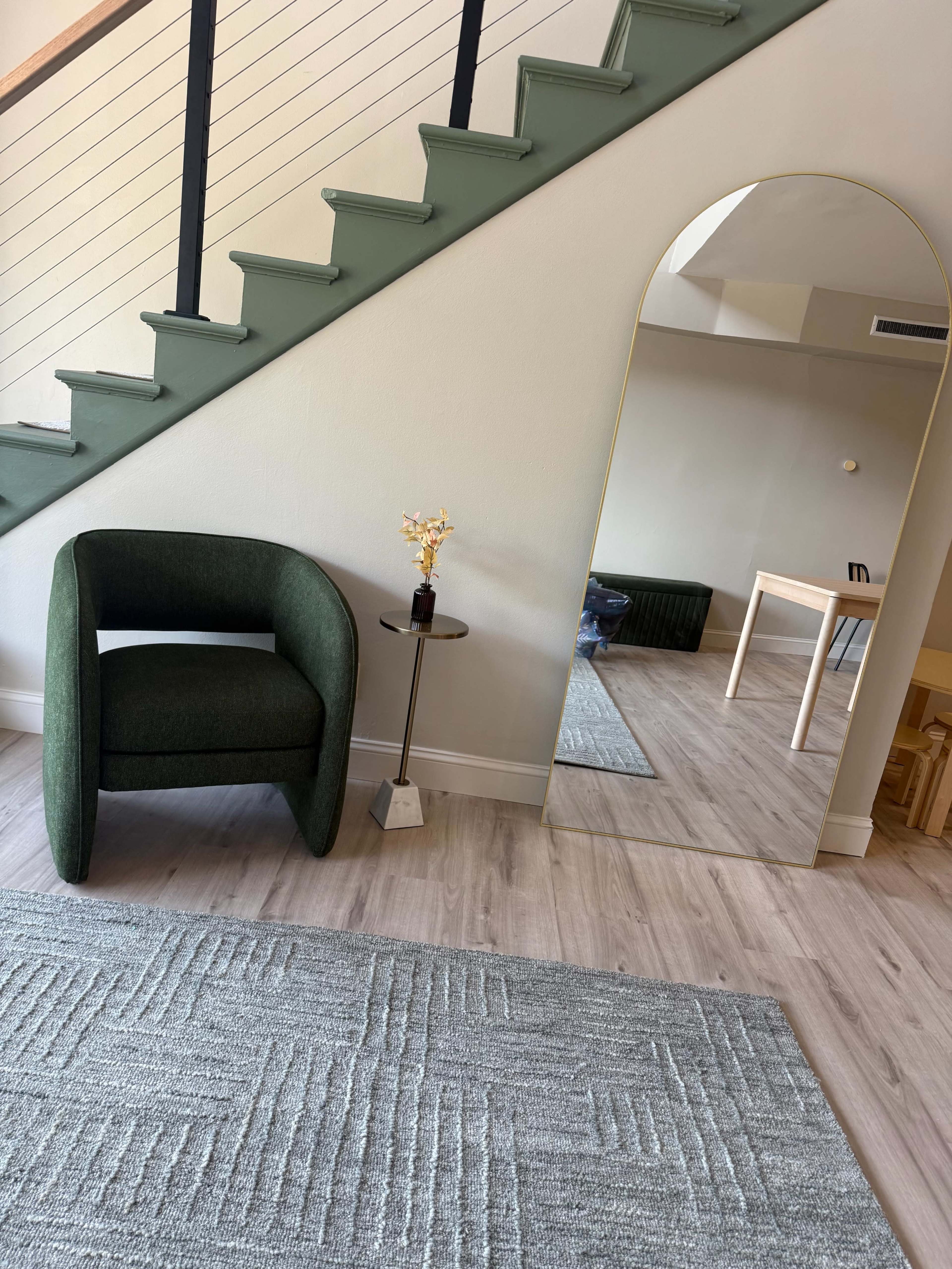 The image shows a green armchair next to a small side table with a decorative flower, facing a tall mirror and a staircase with a light-colored wall.