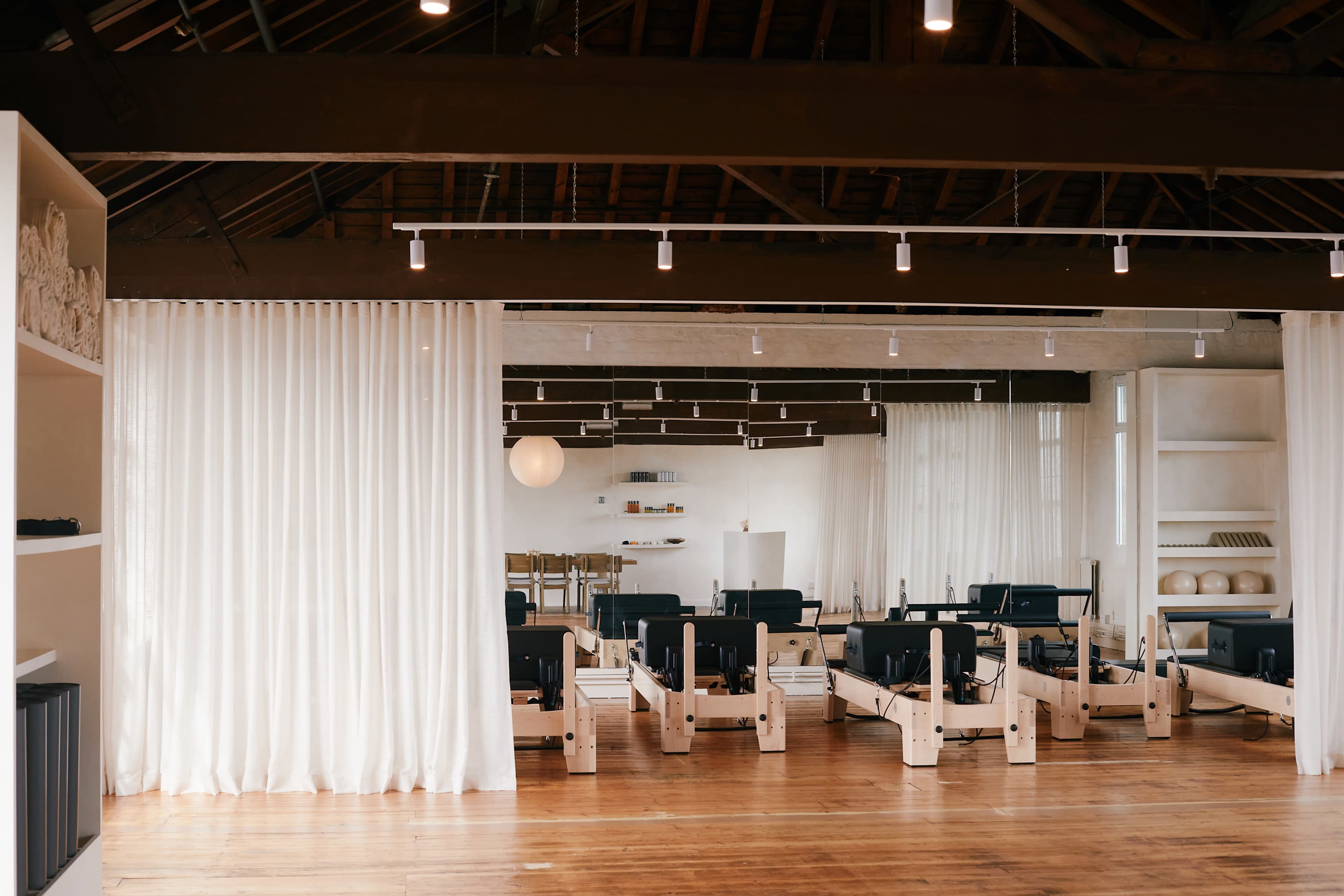 The image shows a spacious studio environment with wooden flooring, featuring several reformer Pilates machines arranged neatly in rows, separated by sheer white curtains.