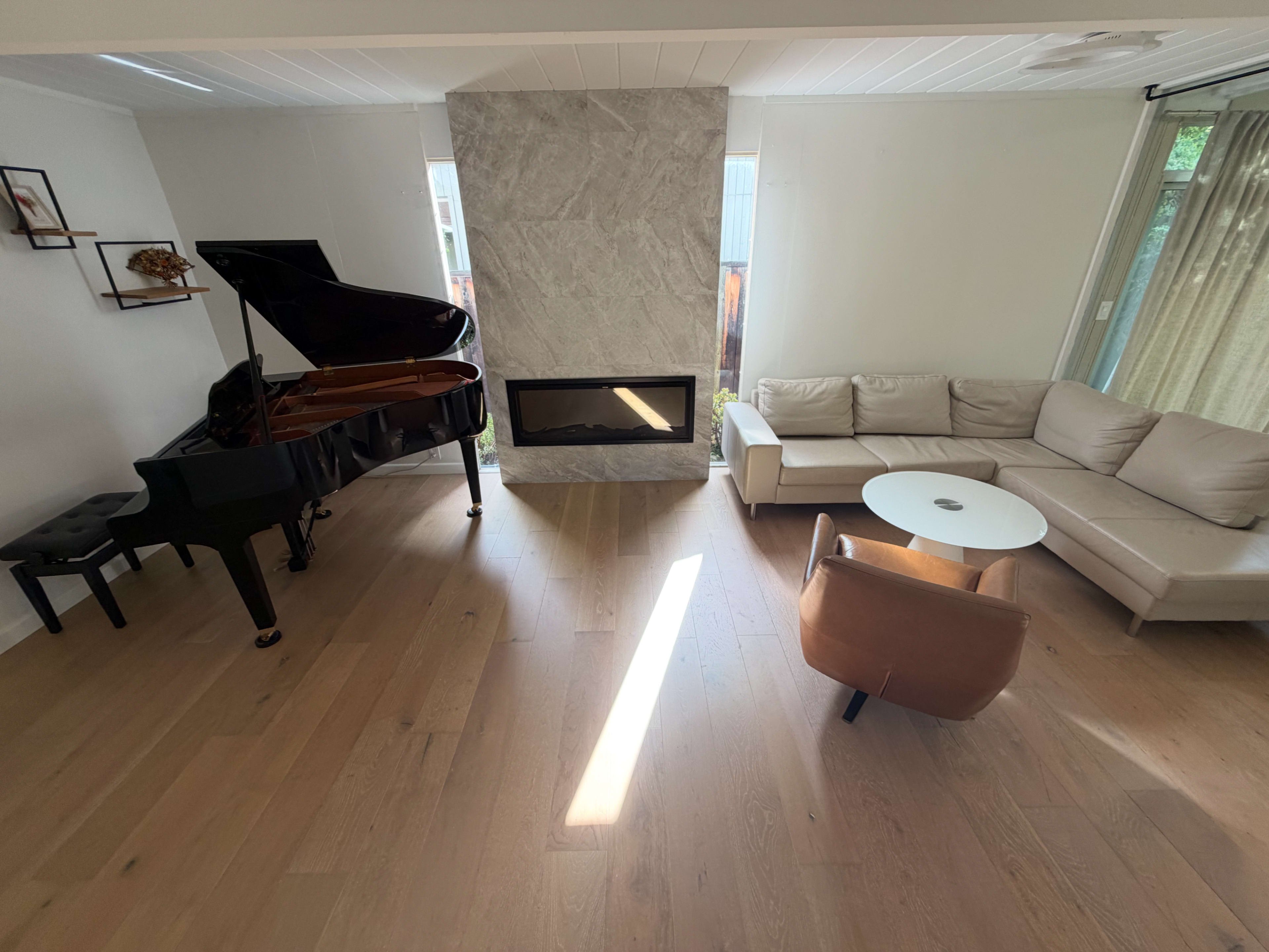 The image shows a modern living room featuring a grand piano, a beige sectional sofa, a small round coffee table, and a stone fireplace.