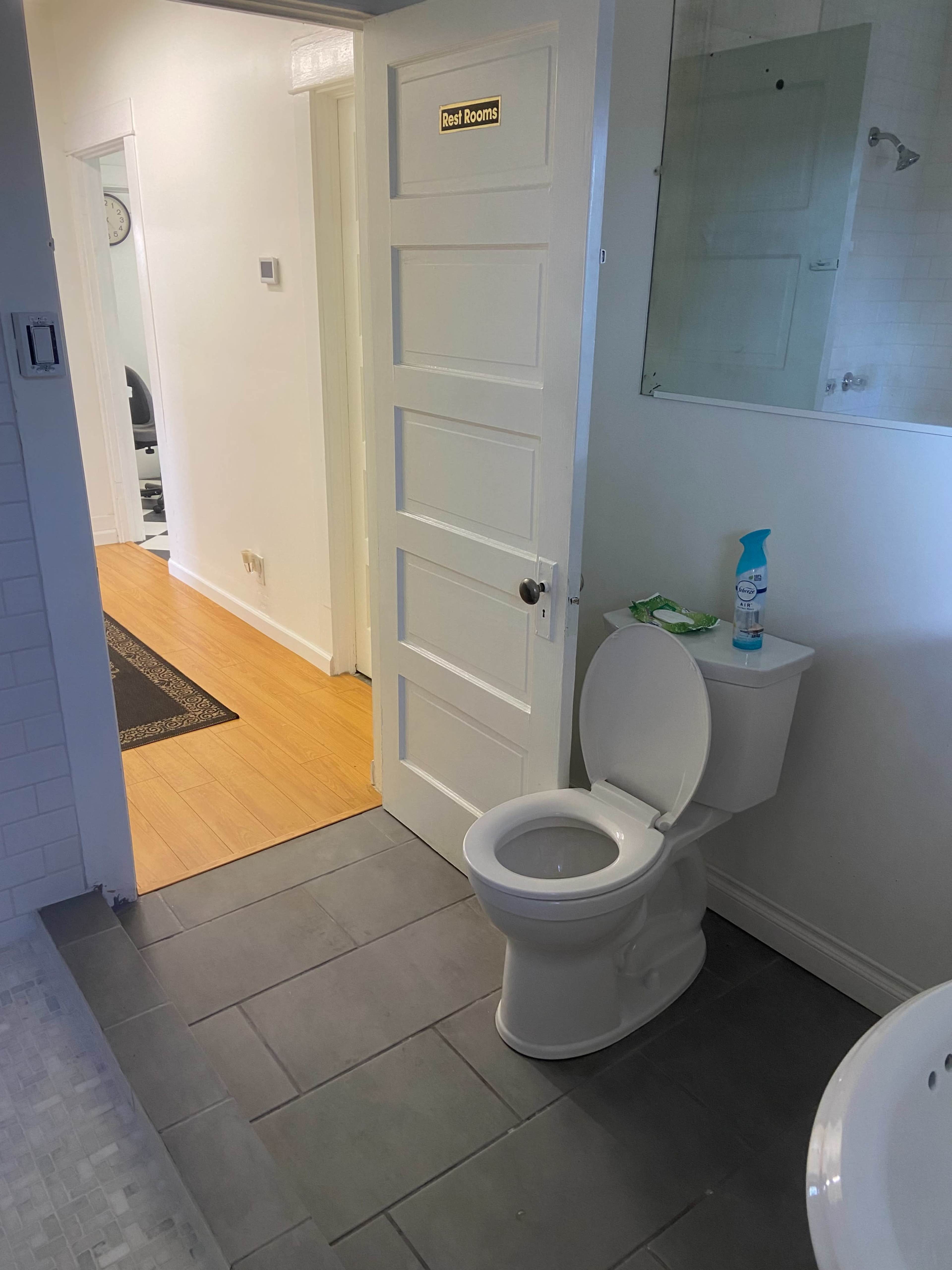 The image shows a bathroom with a white toilet, a door labeled "Rest Rooms," and a glimpse of a hallway with wooden flooring.