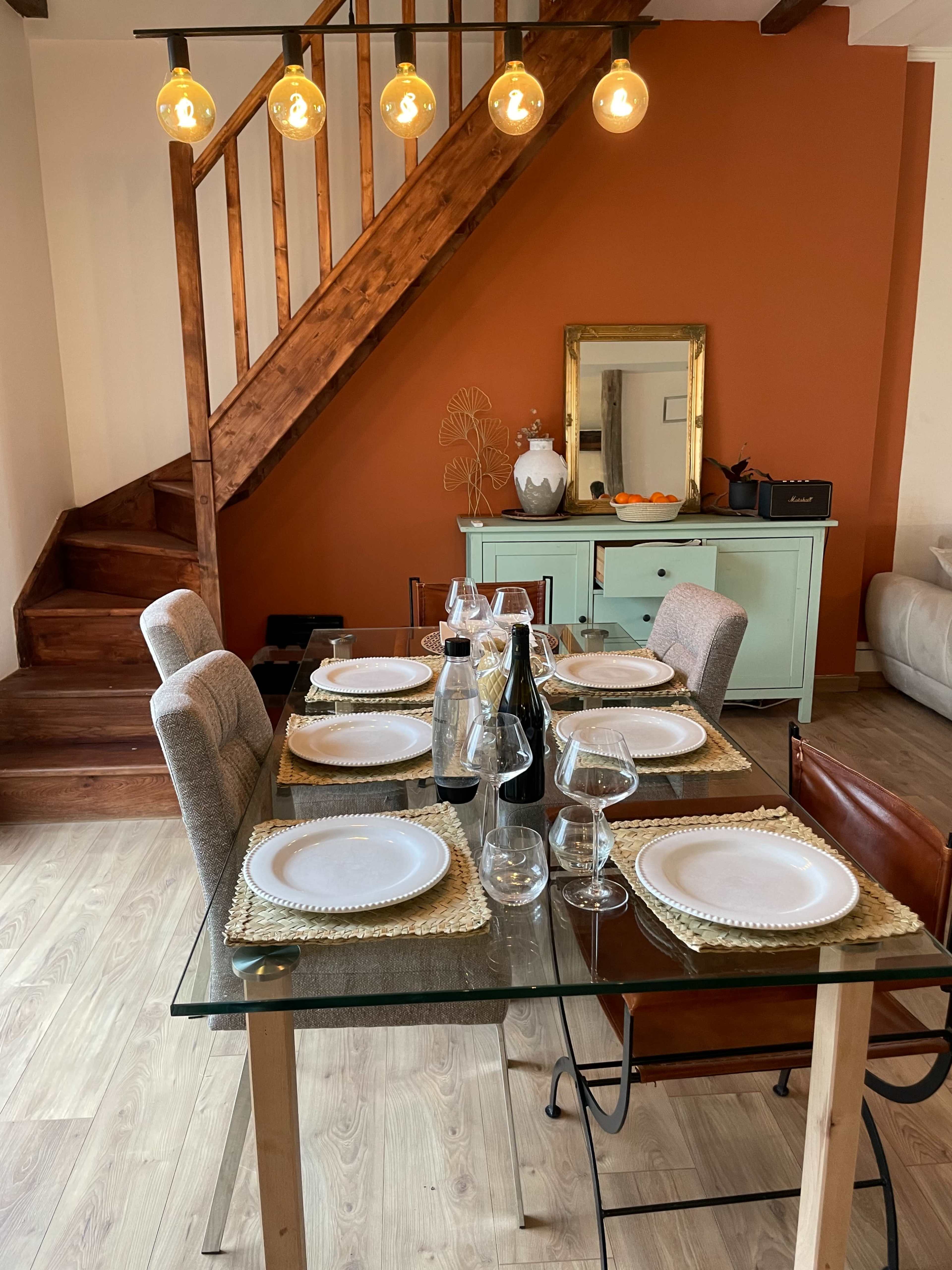 A dining table is set with plates and glassware, positioned near a staircase and a sideboard against an orange wall.