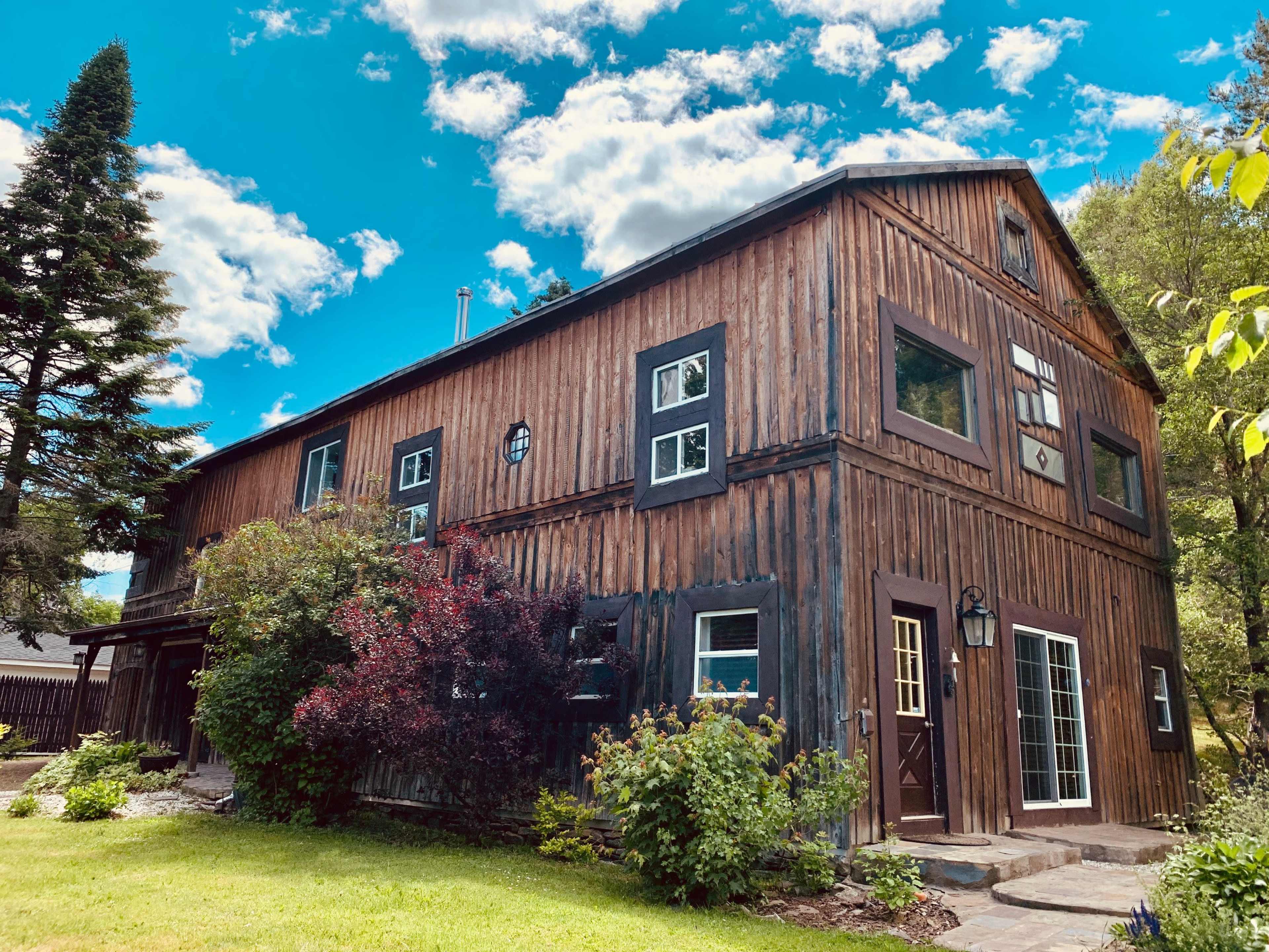 A wooden two-story house with multiple windows and a green lawn is surrounded by trees and shrubs under a blue sky with clouds.