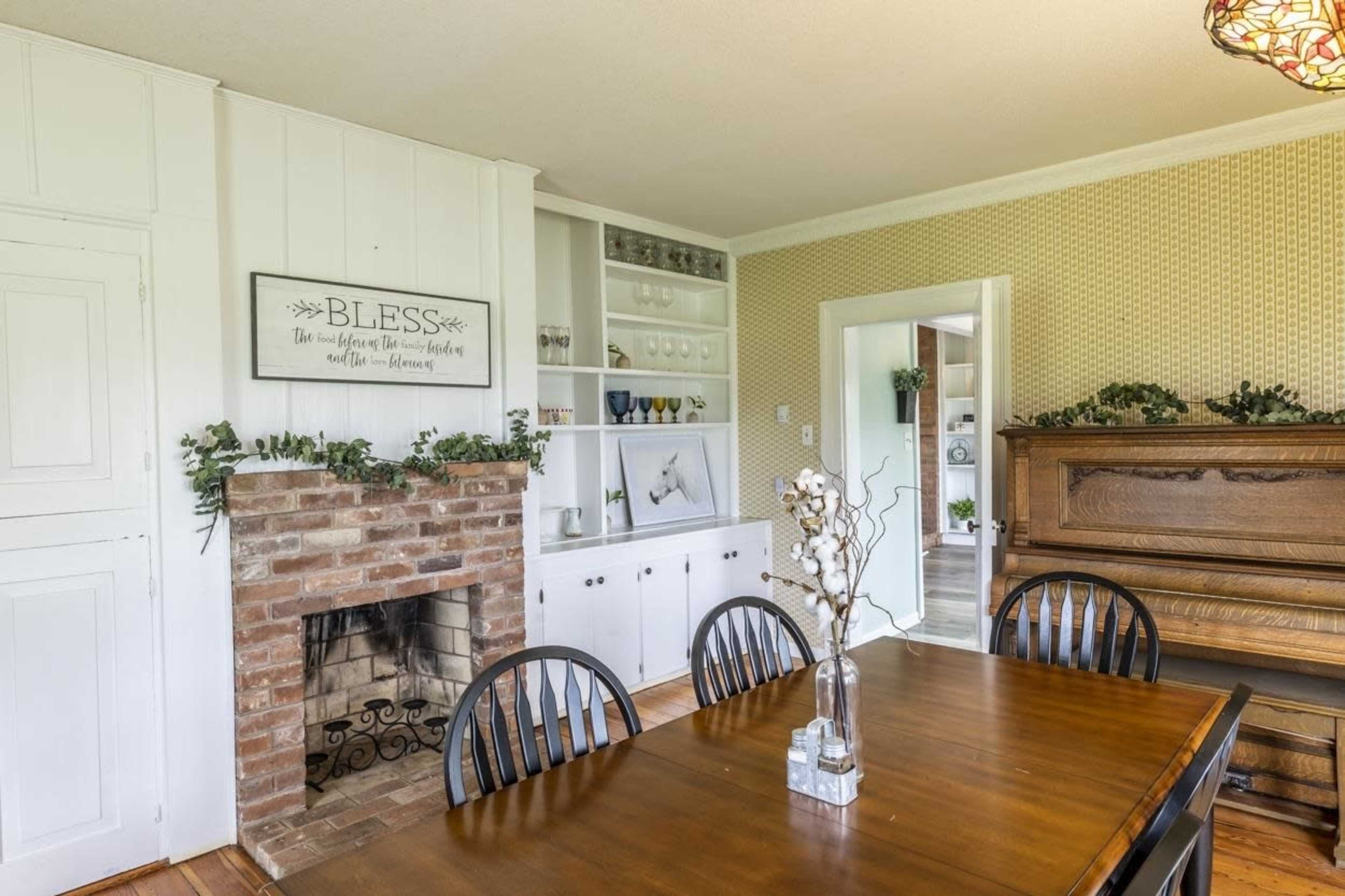A dining room features a wooden table surrounded by black chairs, with a brick fireplace and an upright piano in the background.