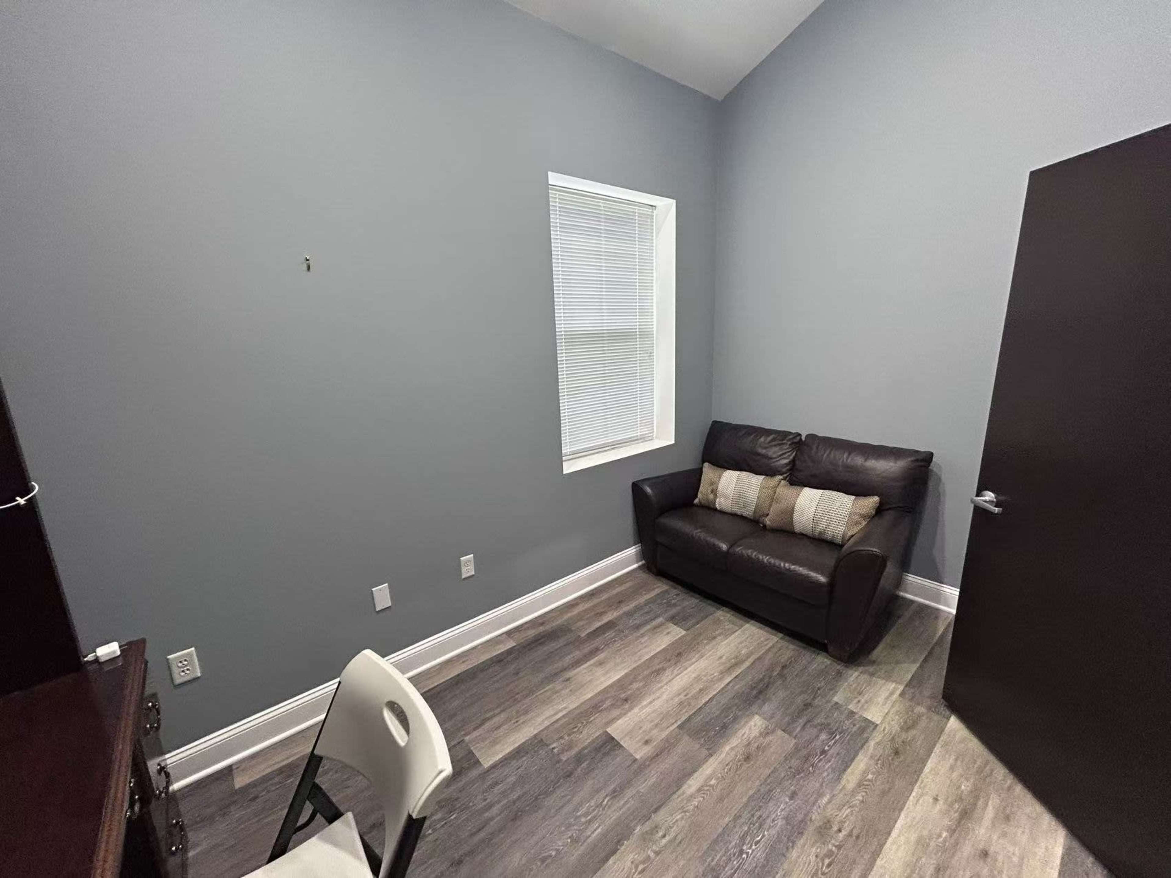 The image shows a small room with grey walls, a brown couch, a chair, and a door leading to the outside.