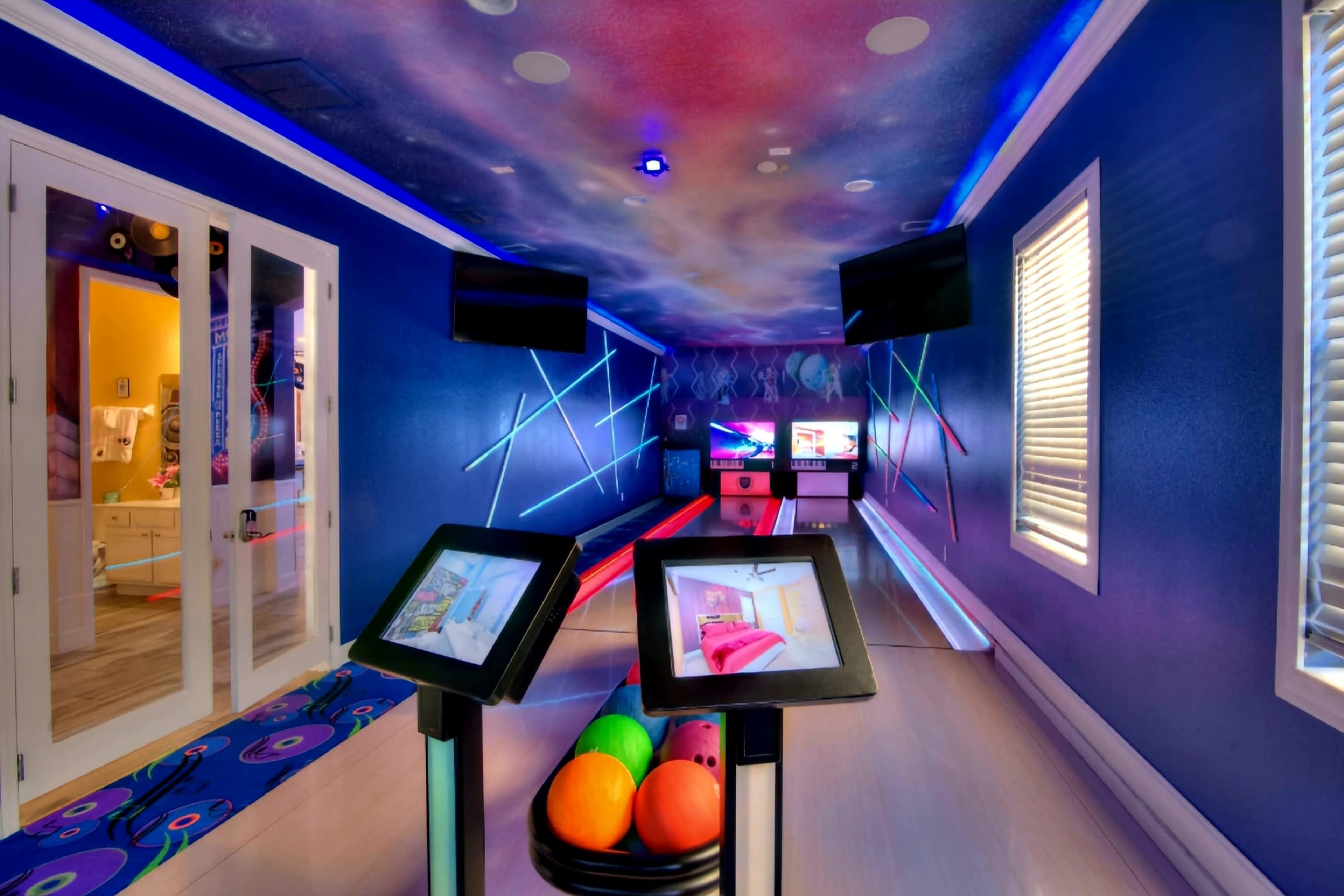 A modern bowling alley with two screens, vibrant lighting, and a set of bowling balls resting on a rack.