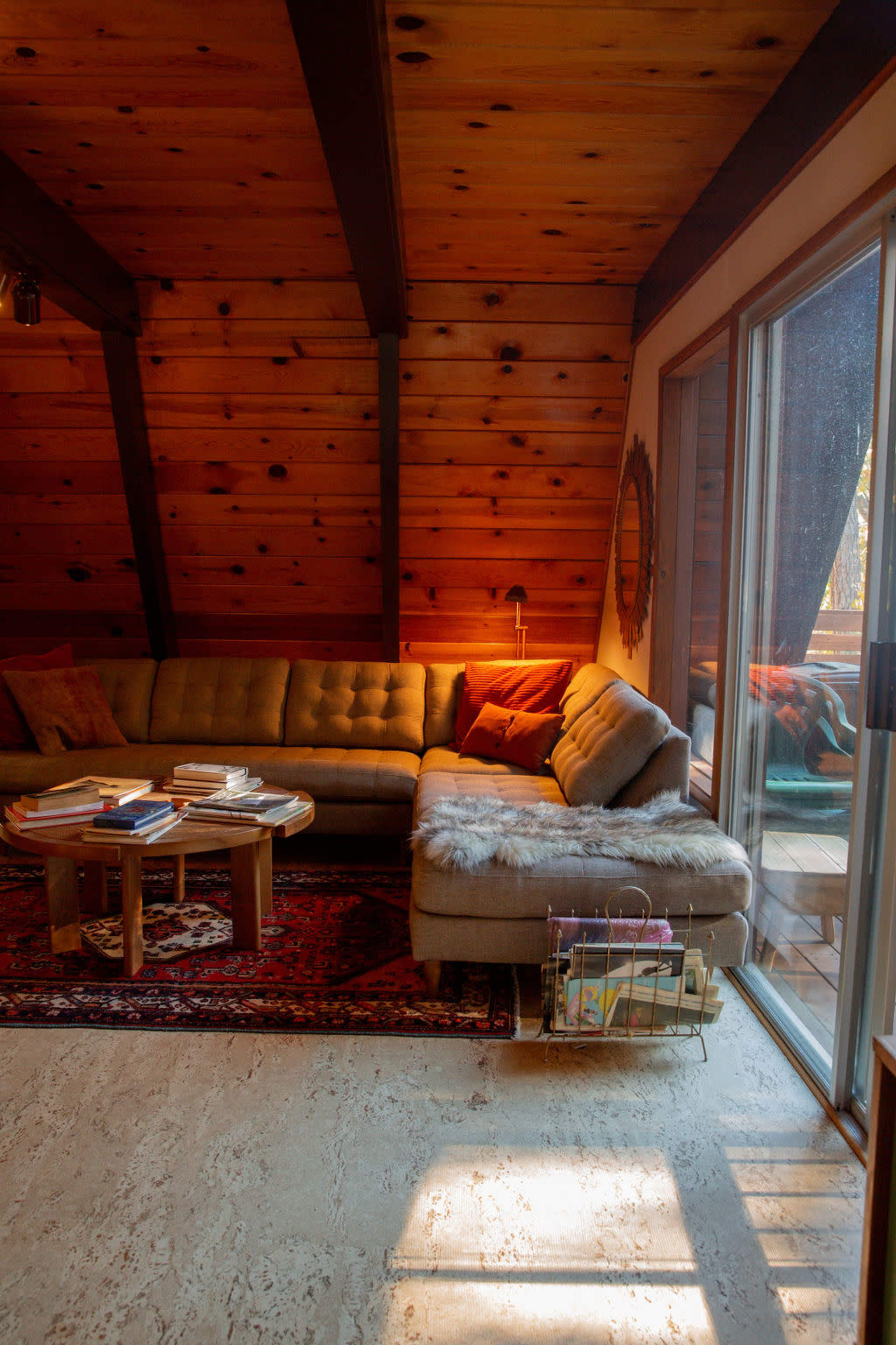 A cozy living room features a large sectional sofa, a wooden coffee table stacked with books, and a floor-to-ceiling window that lets in natural light.