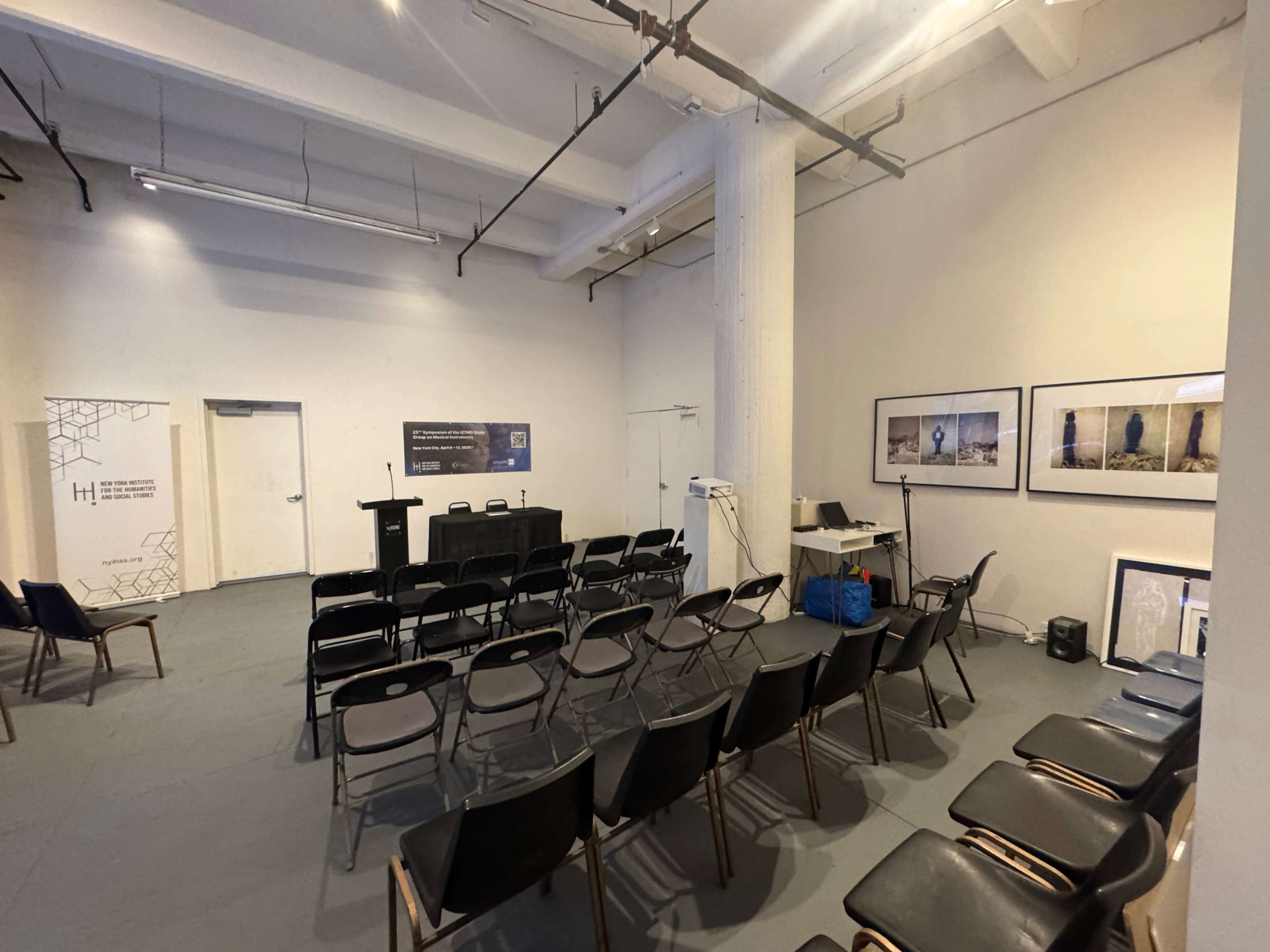The image shows a conference room with rows of black chairs facing a podium and a screen, accompanied by a few framed photographs on the walls.