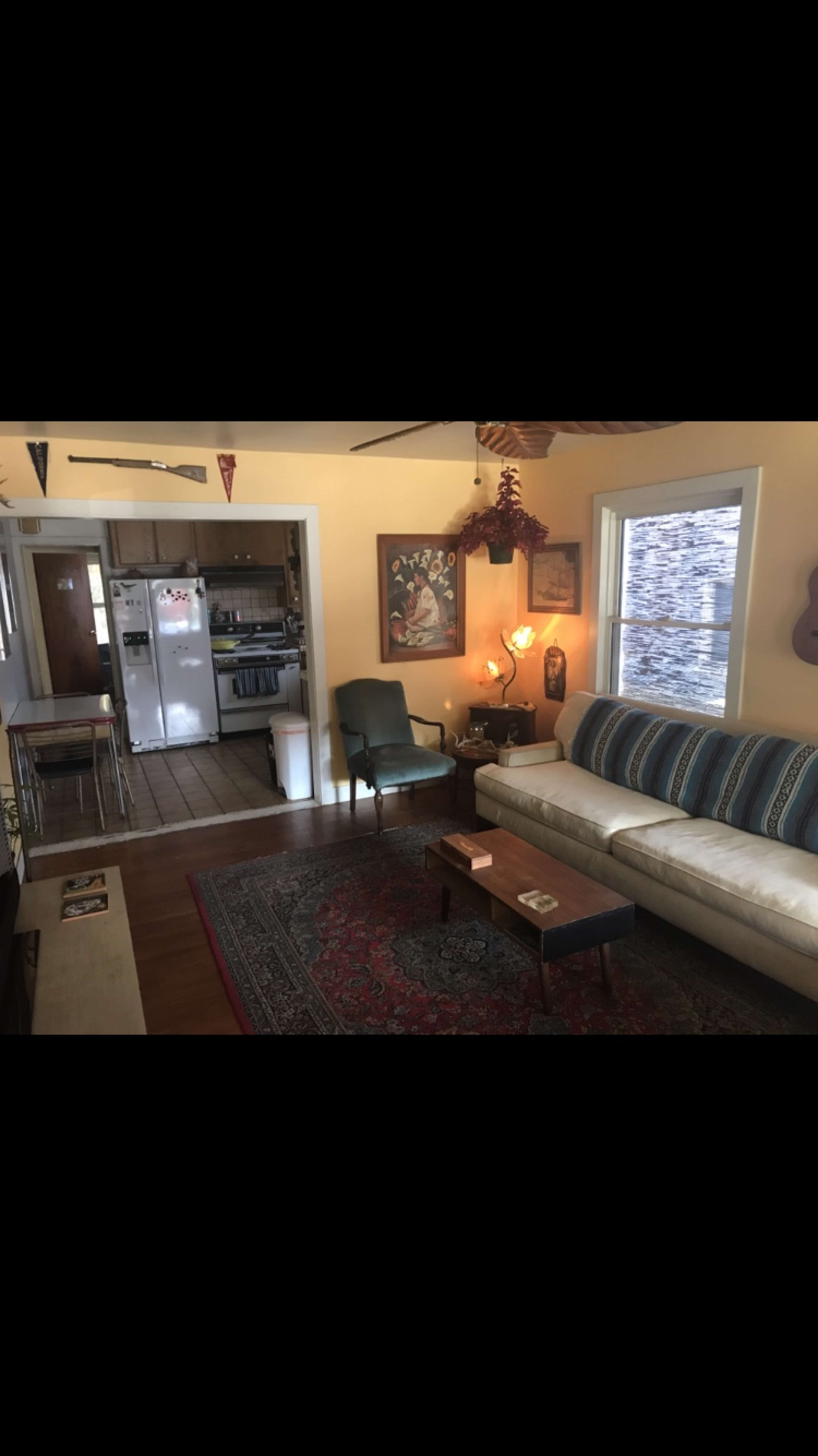 A living room with a sofa, a coffee table, and a green armchair, leading into a kitchen area in the background.