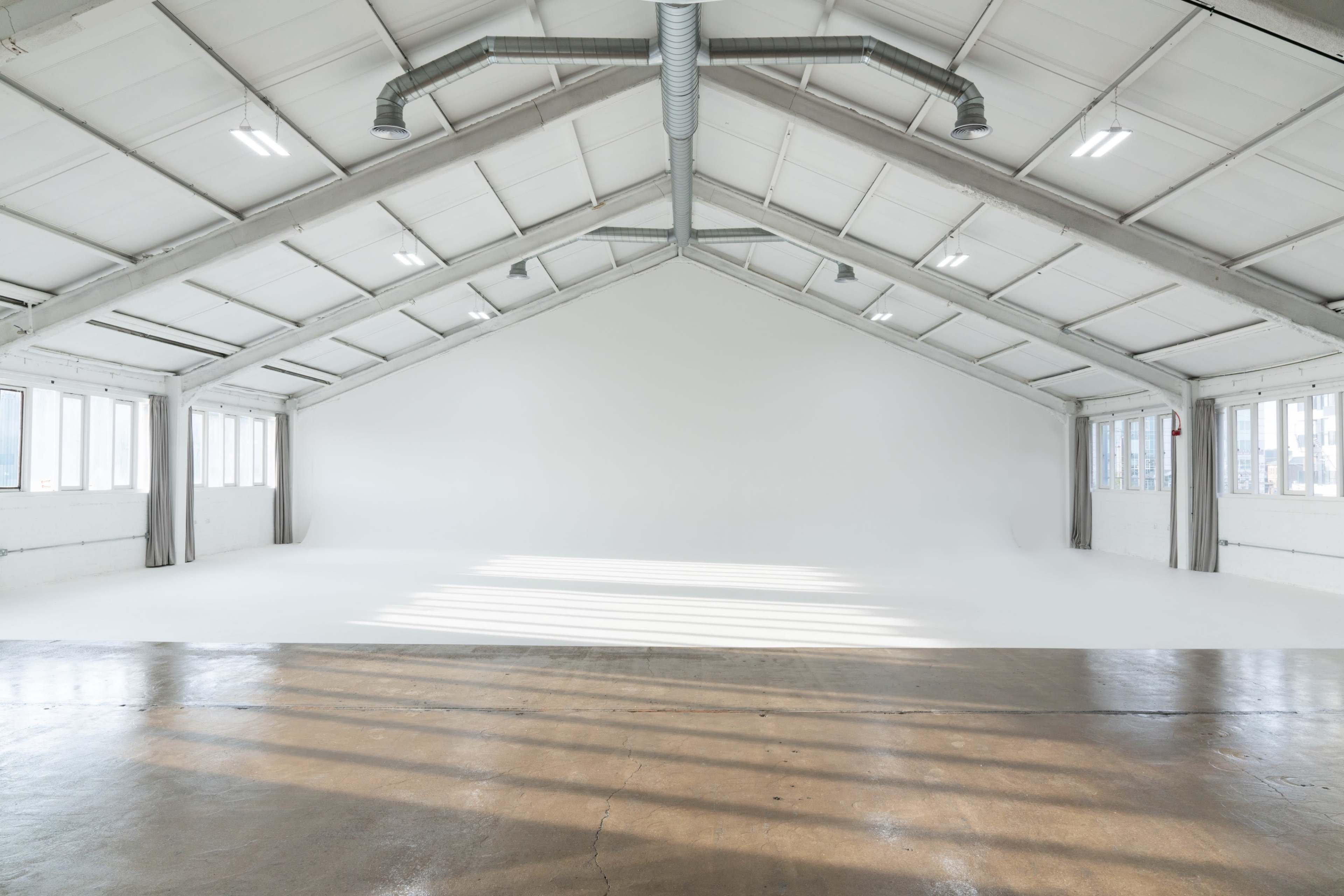 The image shows a spacious, empty industrial-style studio with high ceilings, large windows, and a smooth concrete floor.