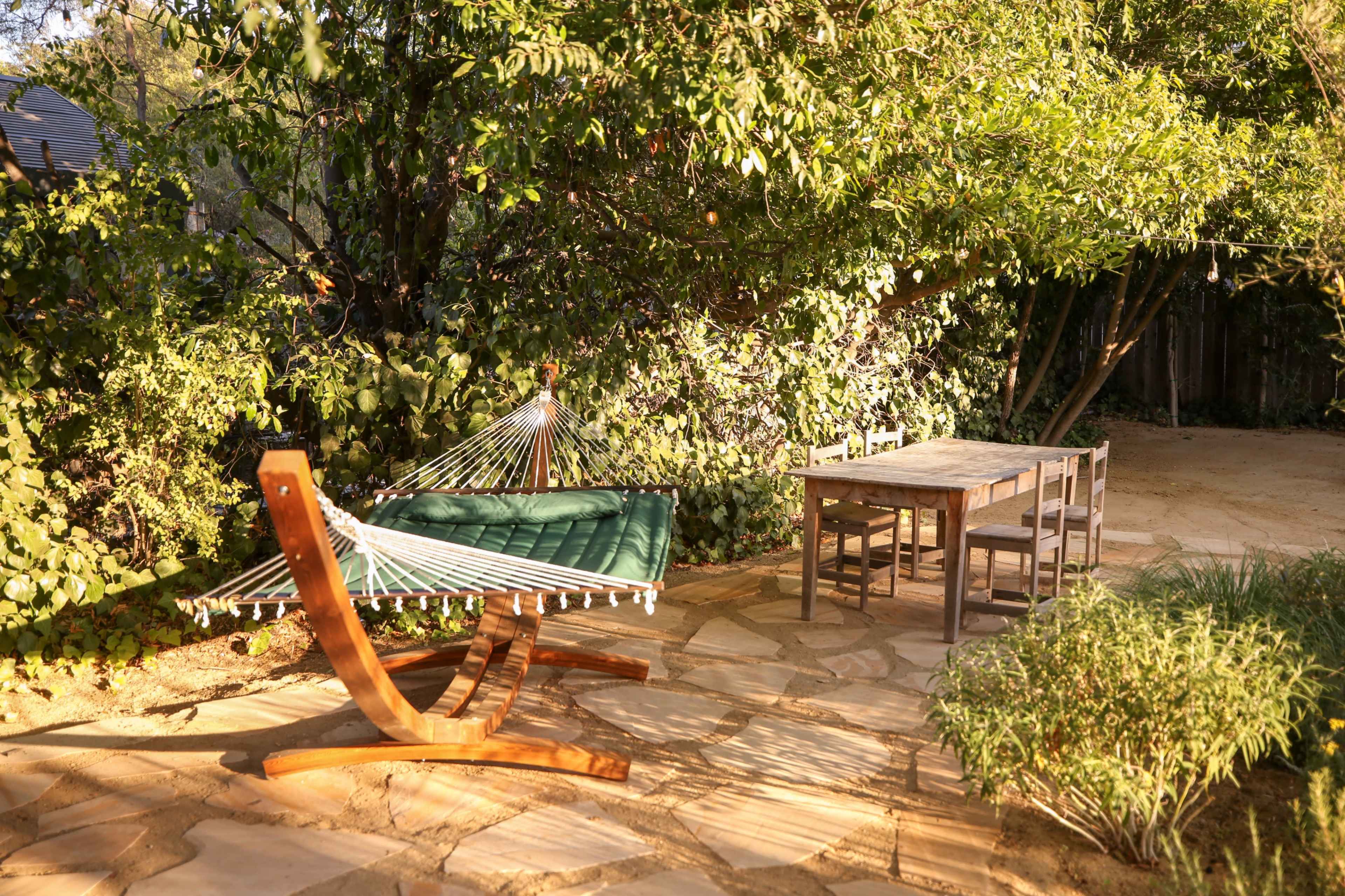 A wooden hammock is positioned next to a small table and chairs on a stone patio surrounded by greenery.