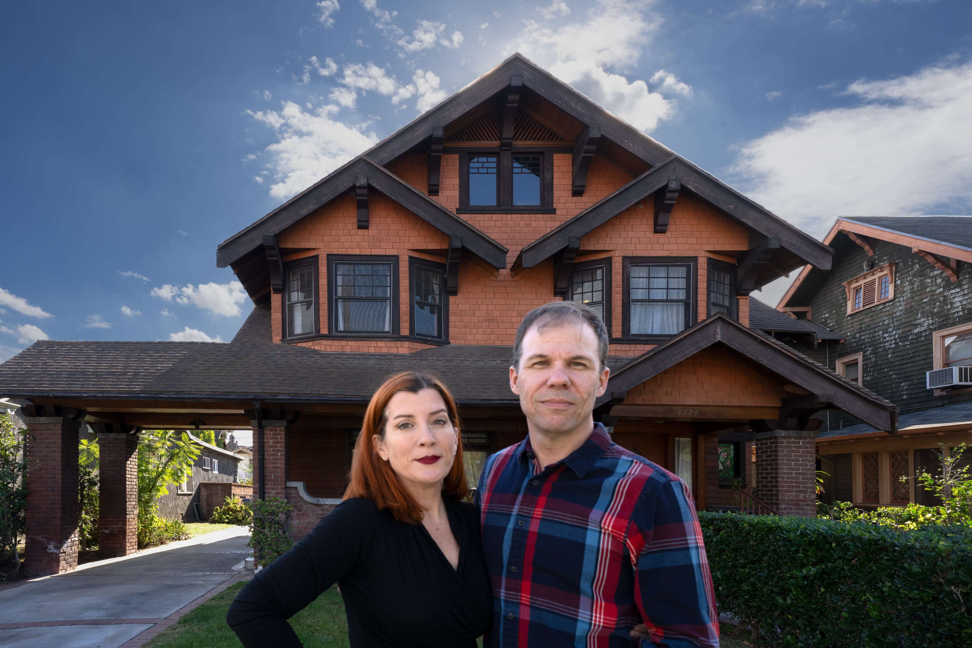A couple poses in front of a large, two-story Craftsman-style house with gabled roofs and a well-kept garden.