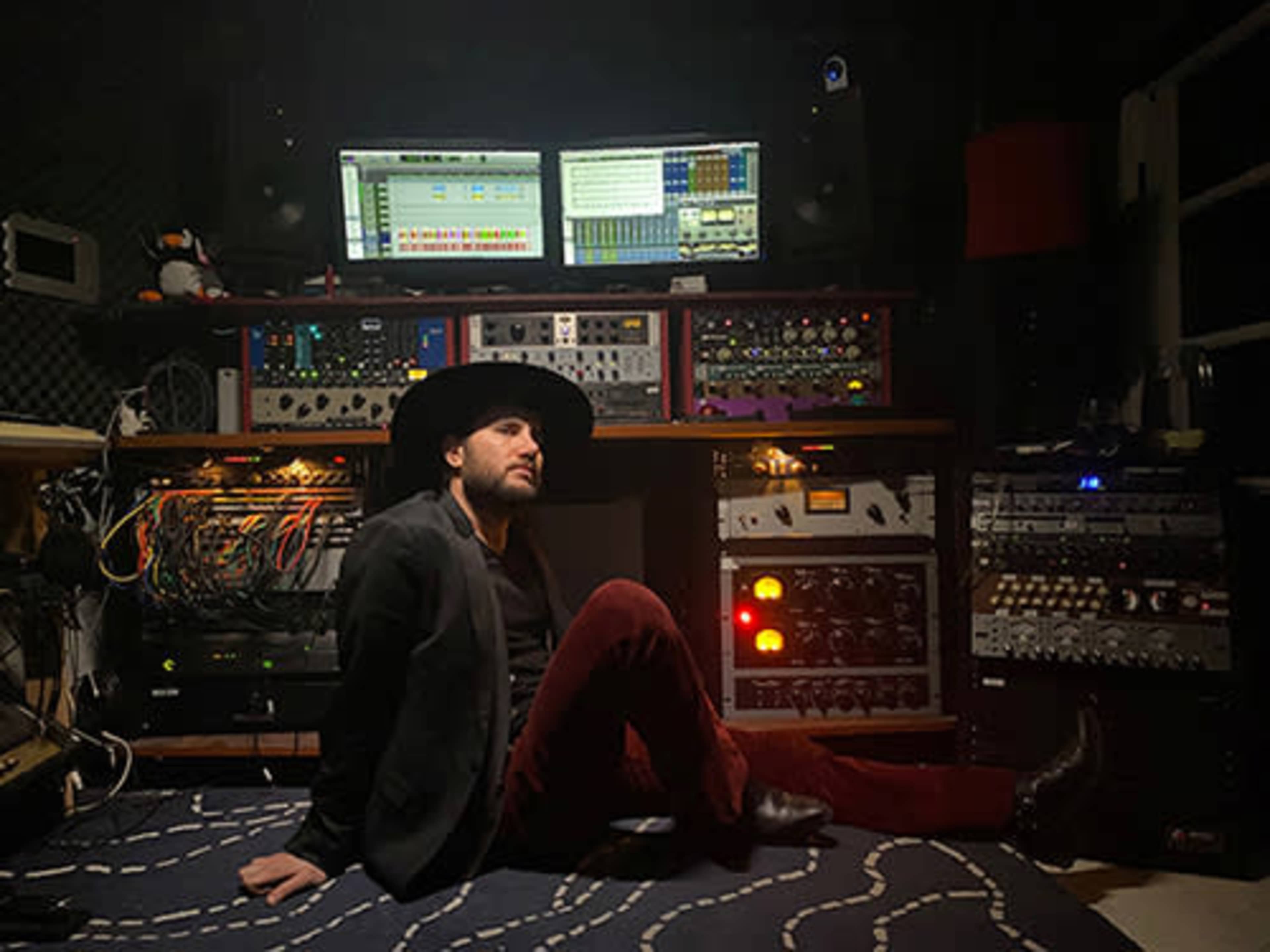 A man sits on the floor of a recording studio surrounded by various audio equipment and two computer screens displaying digital audio tracks.