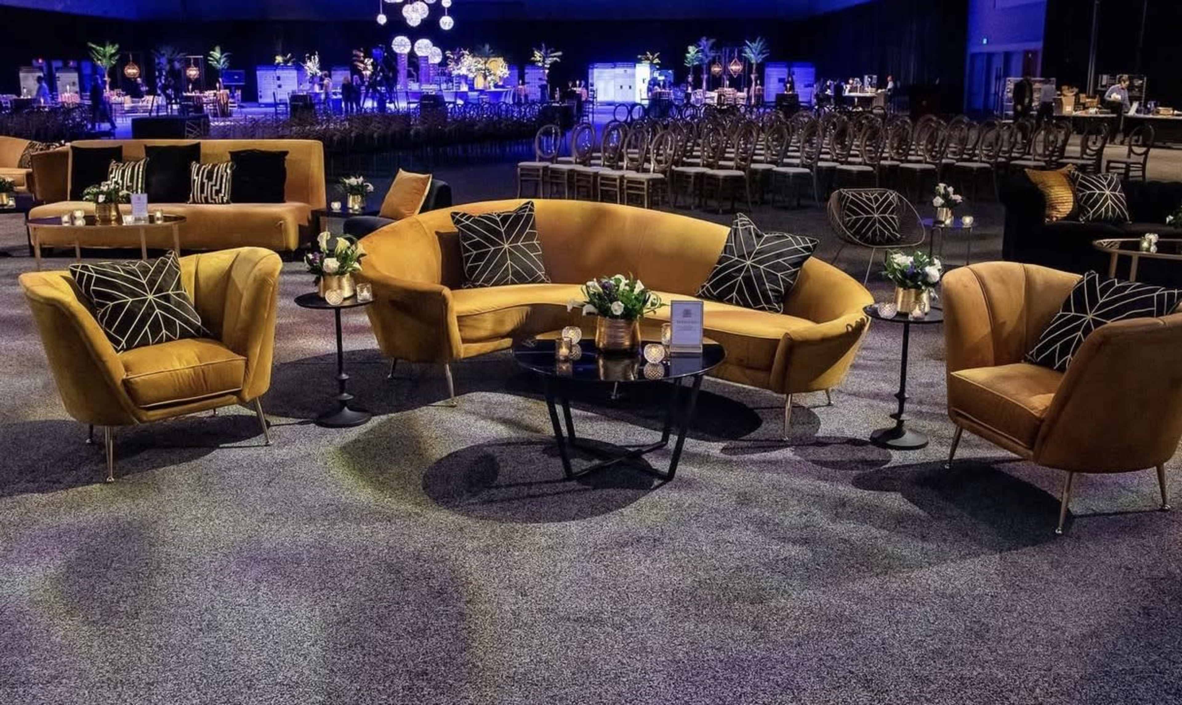 The image shows a stylish seating area featuring yellow upholstered sofas and armchairs arranged around a black coffee table in a spacious event venue.