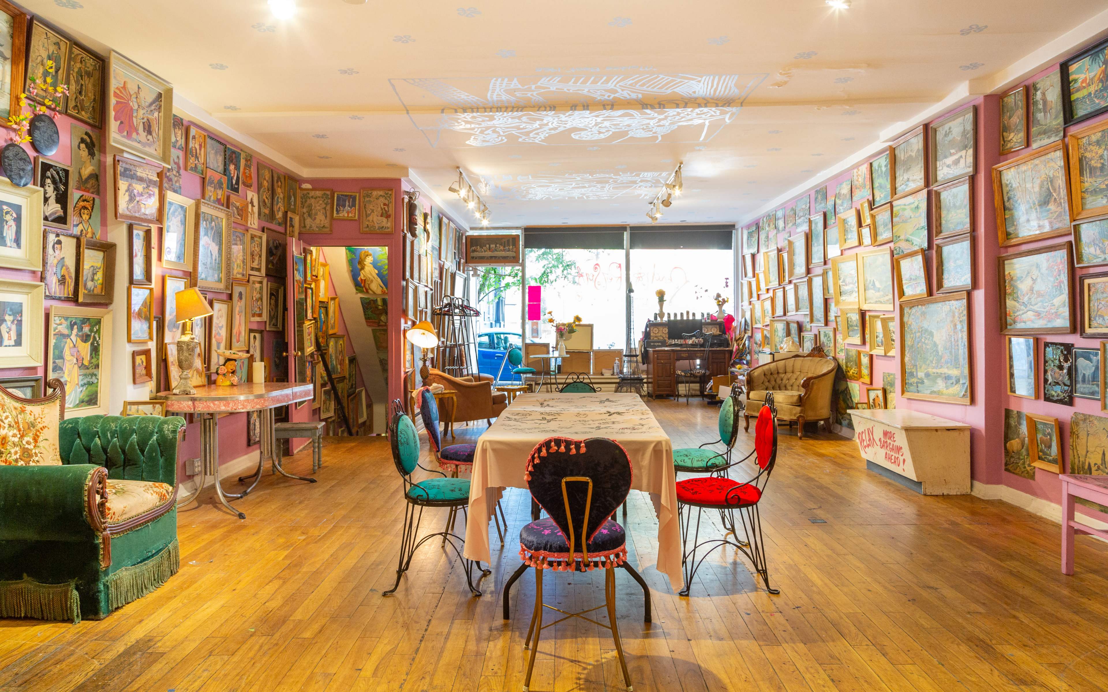The image depicts a colorful art-filled room with a long dining table in the center, surrounded by various chairs, and walls adorned with framed artworks.
