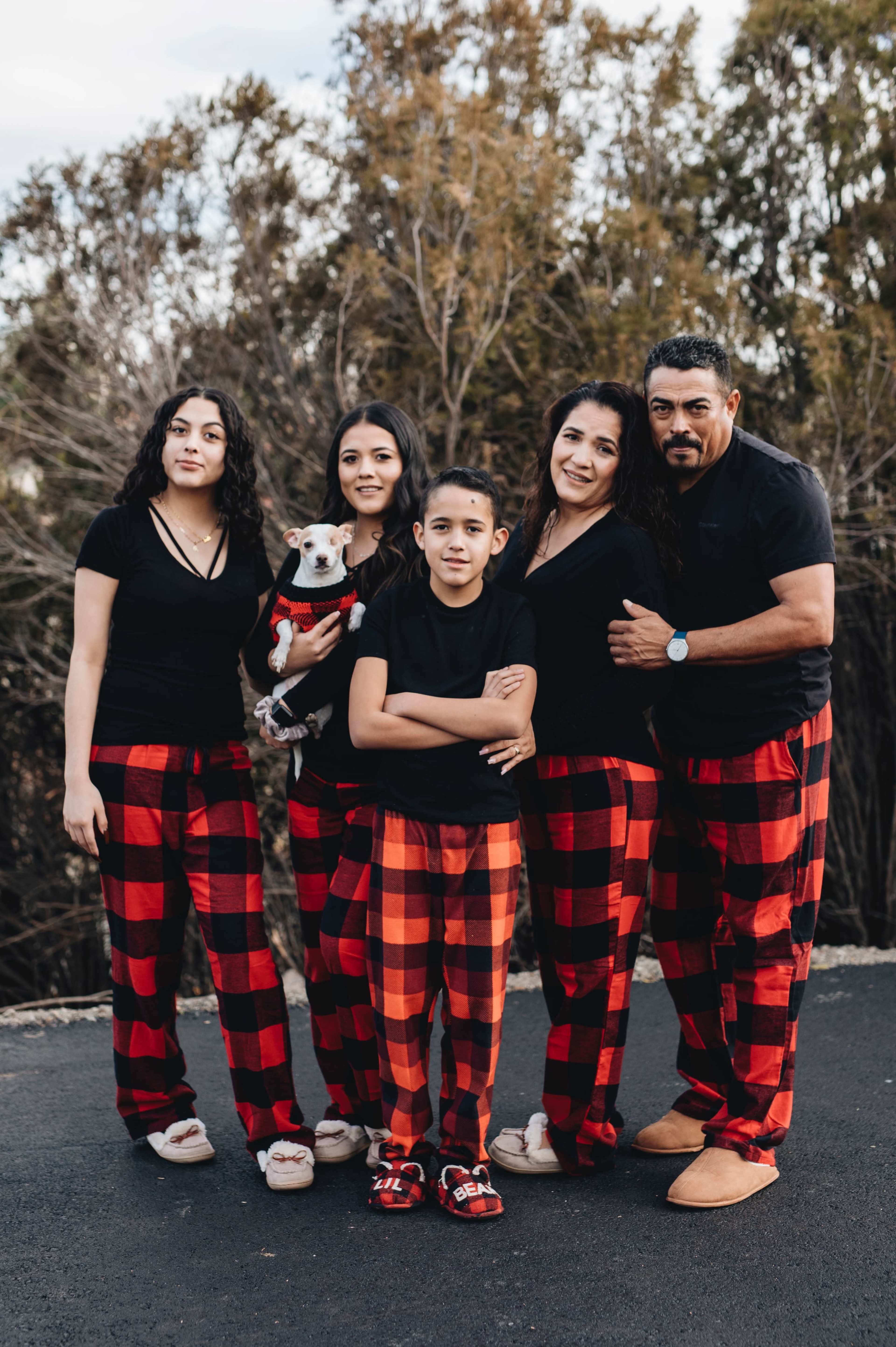 A family of five is standing together outdoors, wearing matching black shirts and red plaid pajamas, with one member holding a small dog.