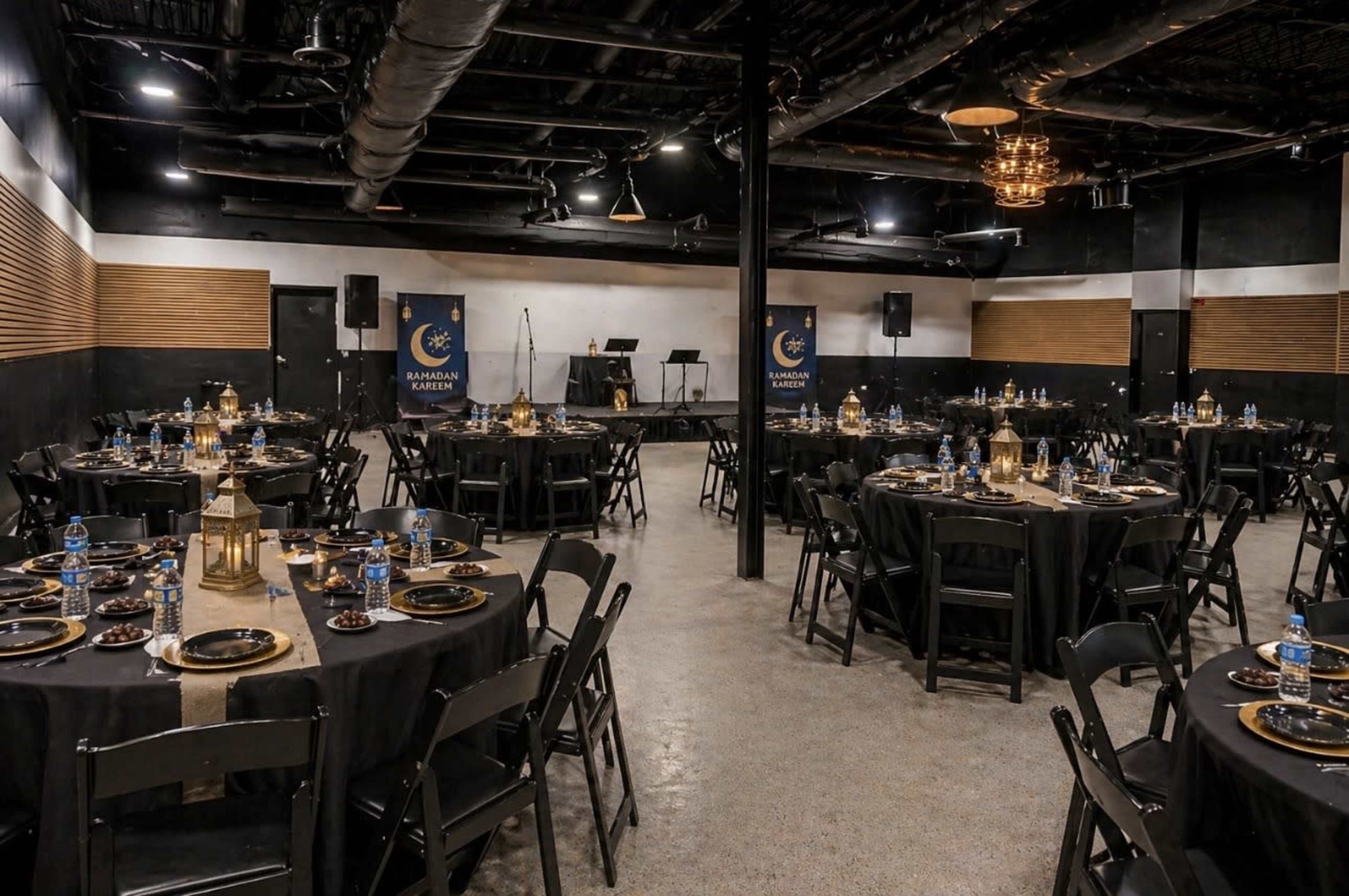 The image shows a large indoor event space set up for a banquet, featuring round black tables arranged with plates, utensils, and gold accents, alongside a stage area with audio equipment.