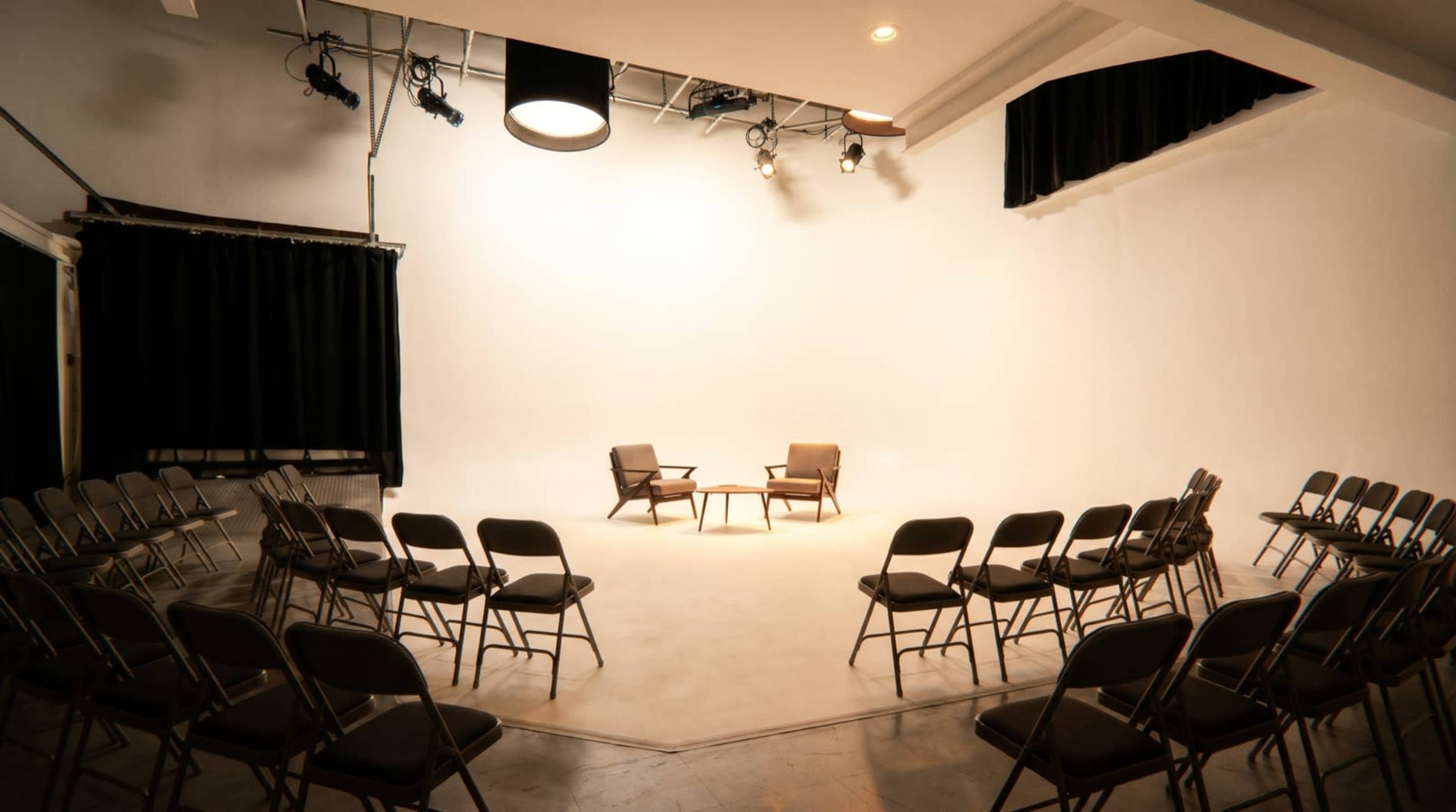 The image shows a minimalistic studio setting with two chairs placed in the center and rows of empty folding chairs arranged around them.