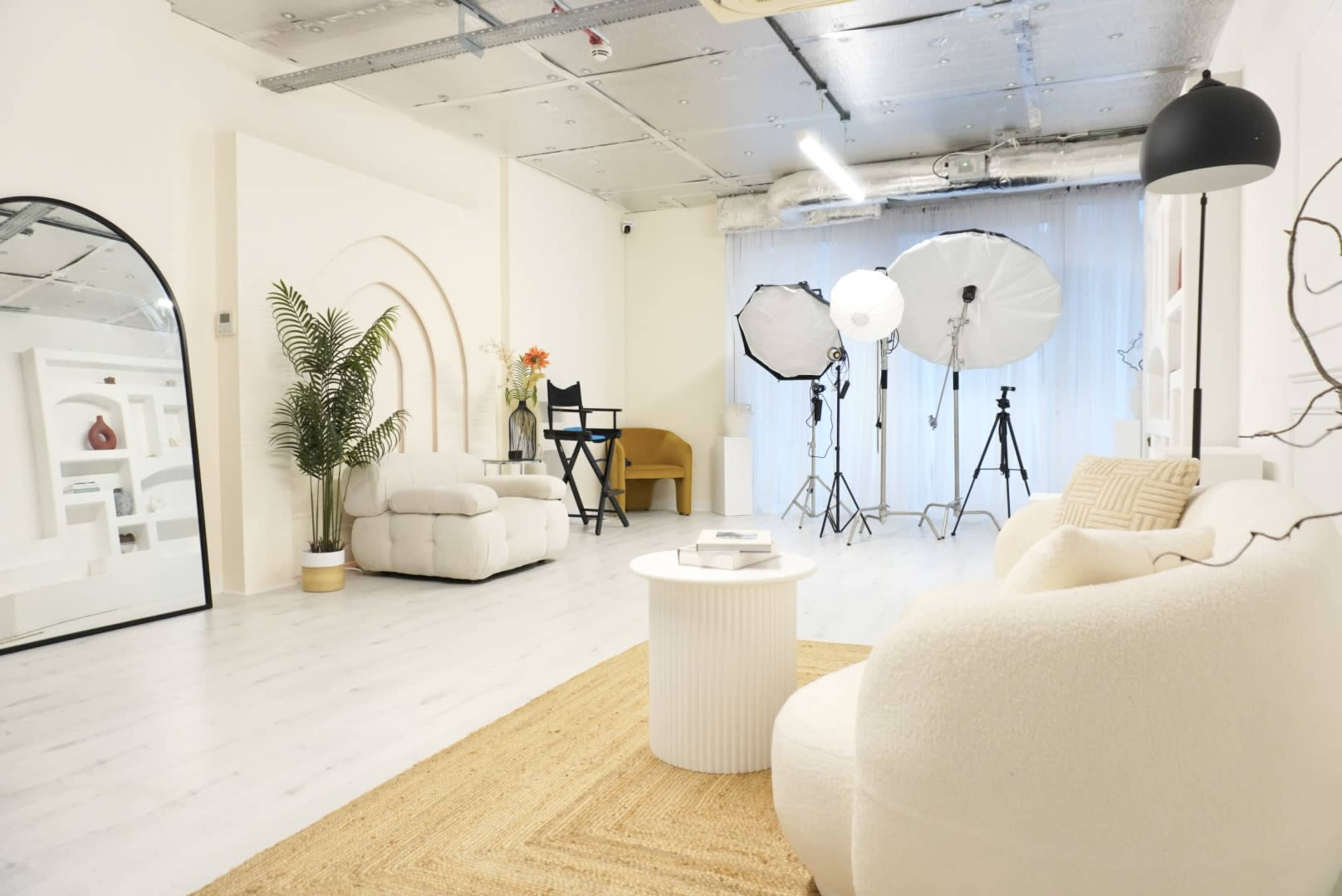 The image shows a bright, modern studio space featuring a large mirror, a cozy seating area with a round table, a plant, and photography equipment including softboxes in the background.