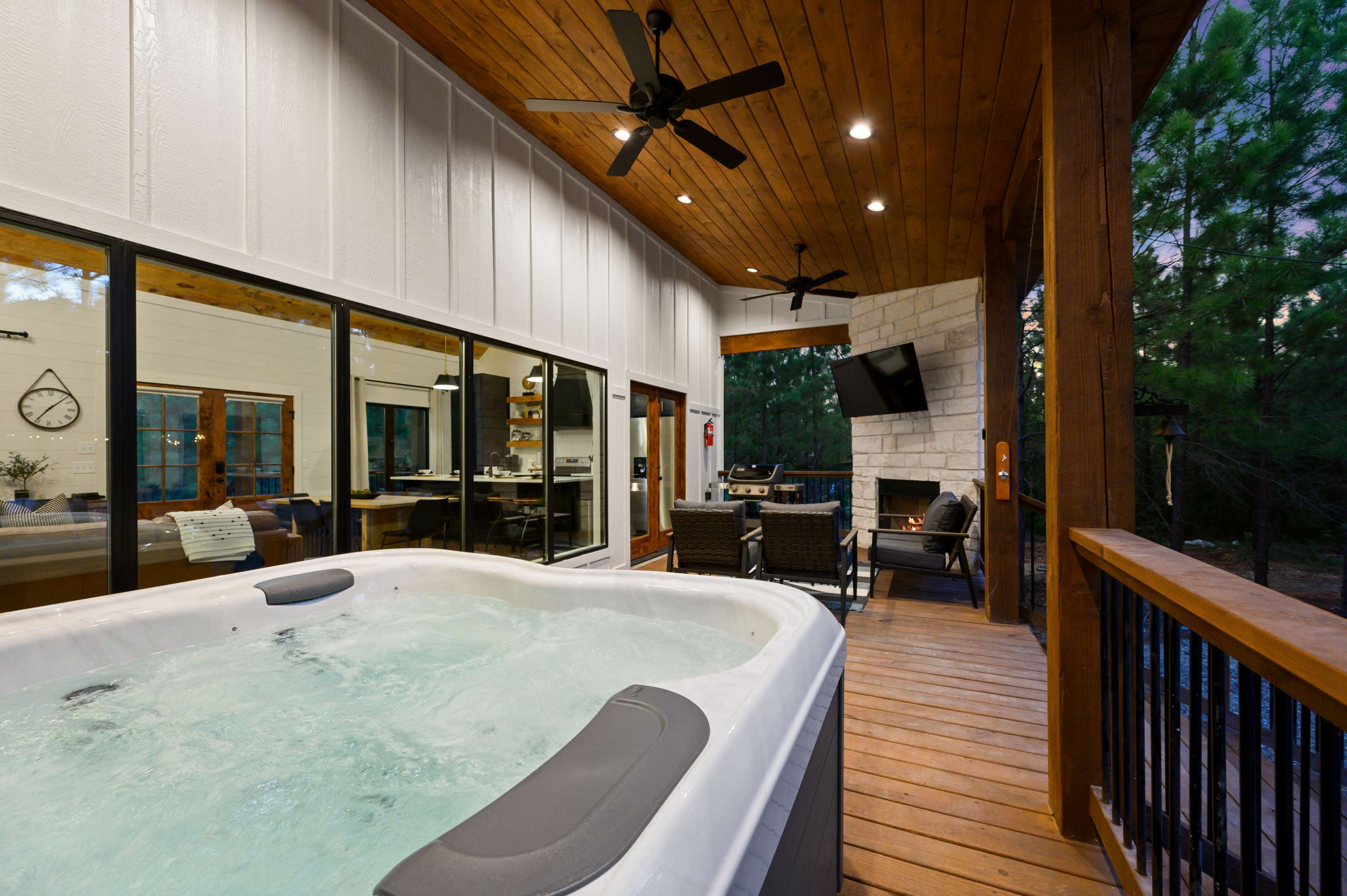 A covered patio with a hot tub, seating area, and large windows overlooking a wooded area.