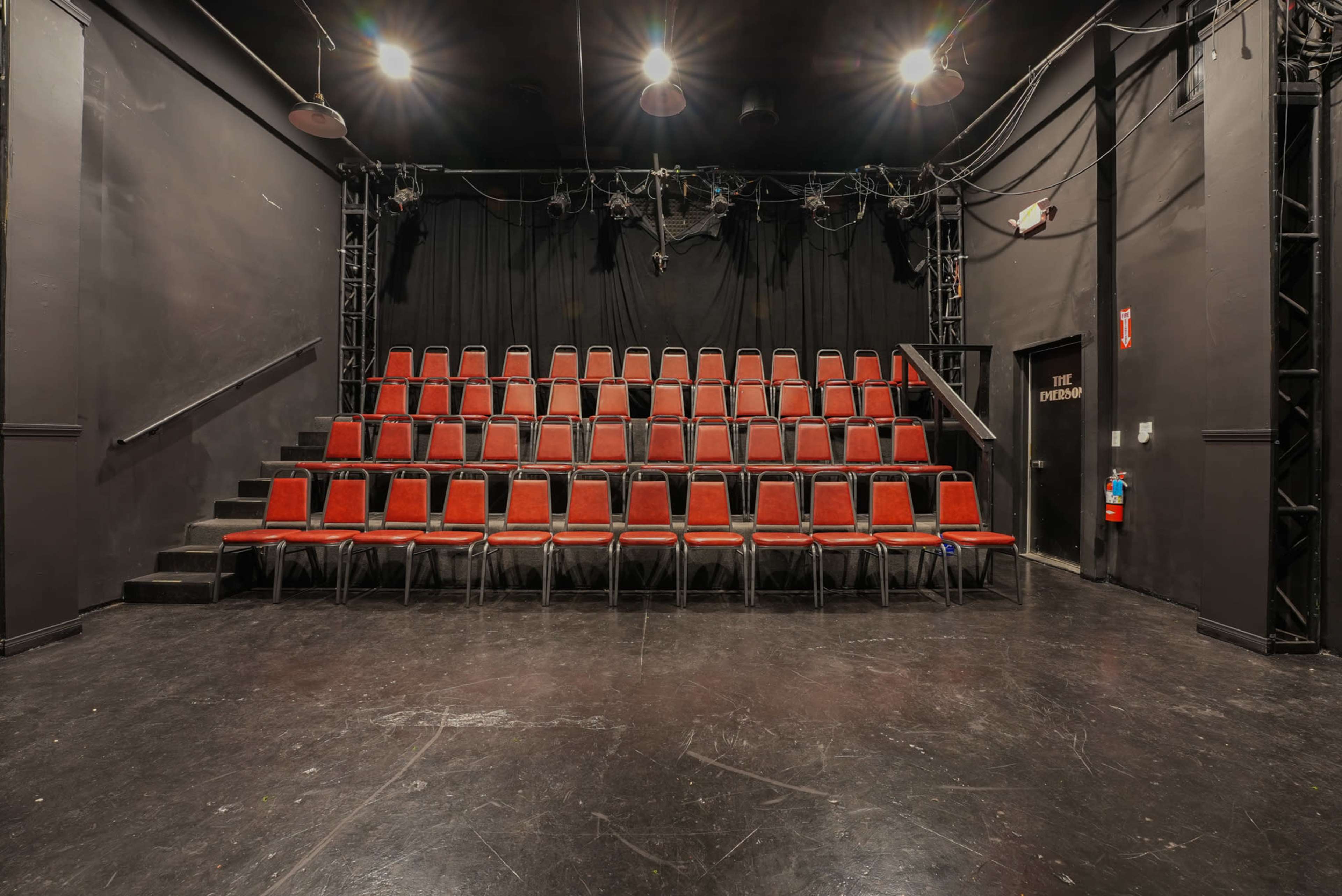 The image shows a dimly lit theater with a set of red chairs arranged in rows facing a black backdrop.