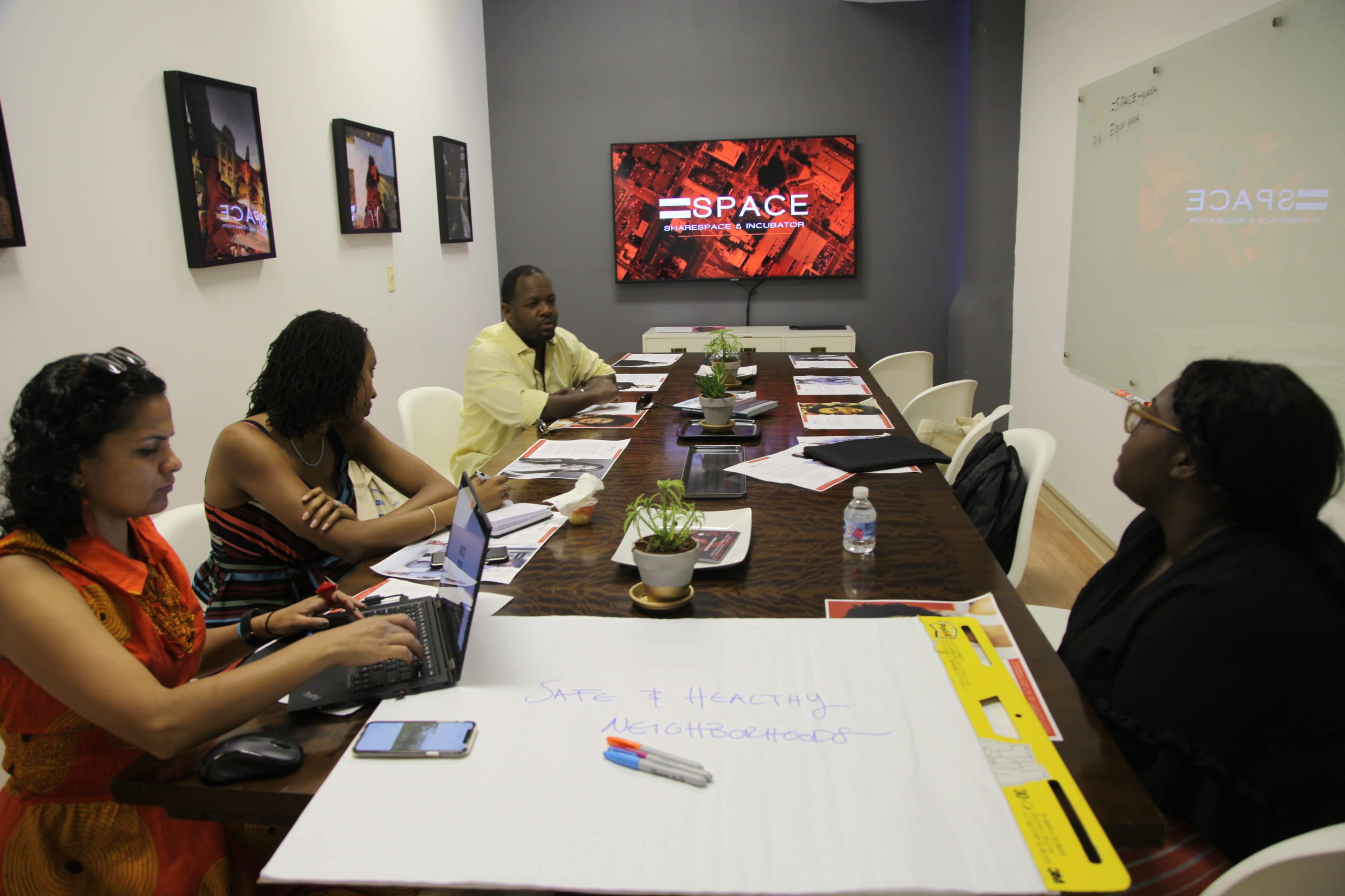 A group of four people is seated around a conference table in a meeting room, with a screen displaying the word "SPACE" in the background.