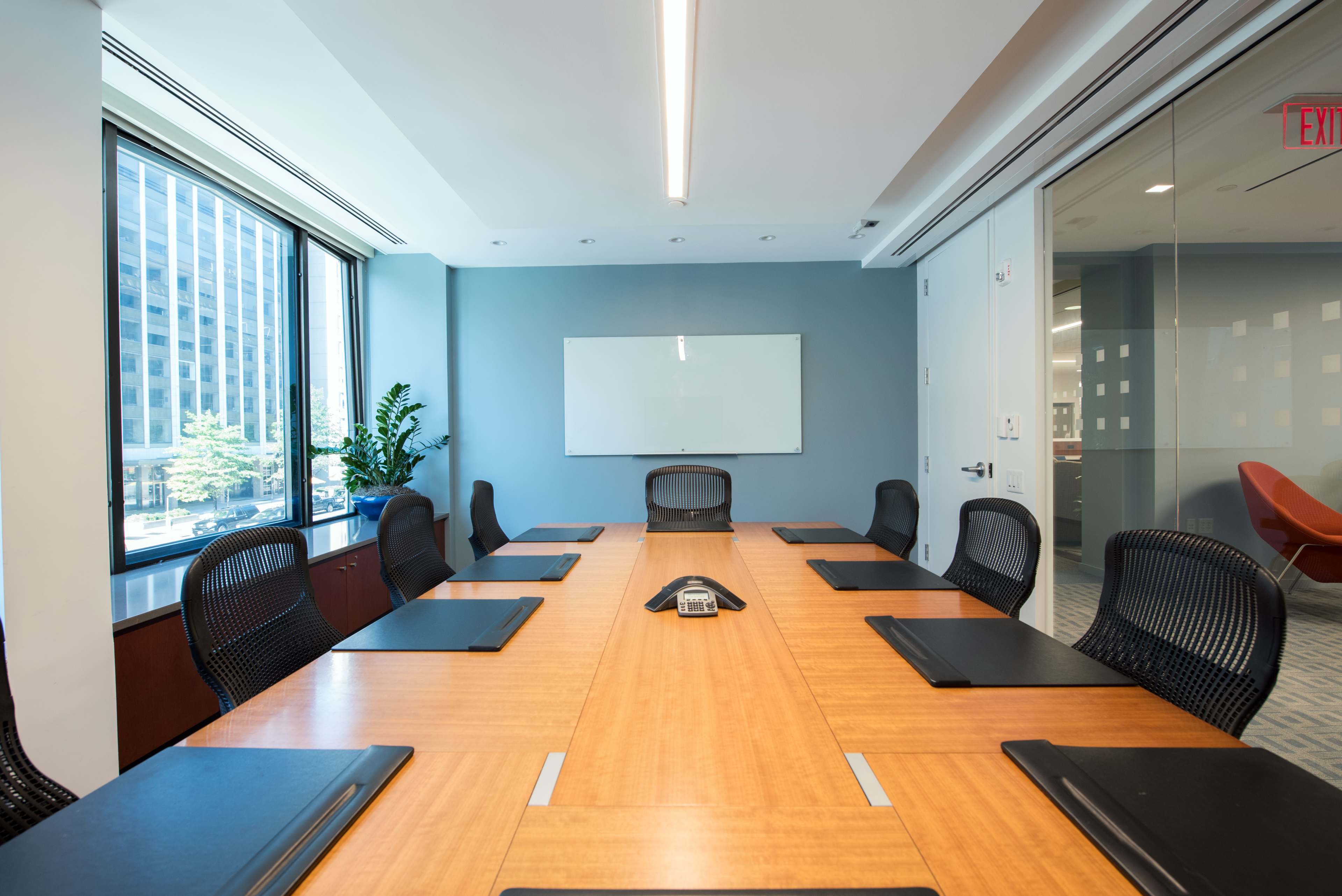 A modern conference room features a long wooden table surrounded by black chairs, with a whiteboard and large windows overlooking a cityscape.