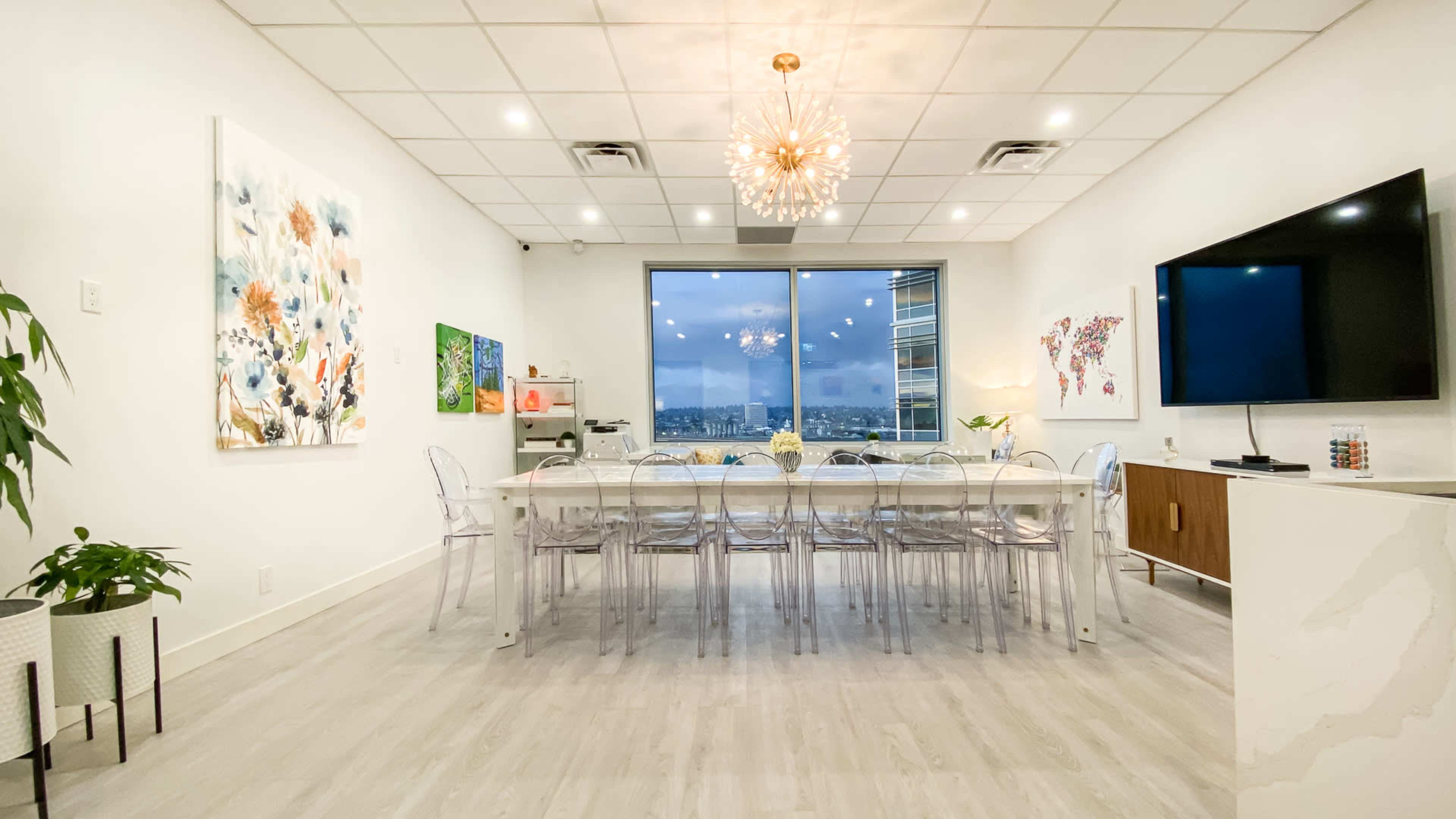 A modern meeting room features a long white table, clear chairs, and large windows with a view of the city skyline.