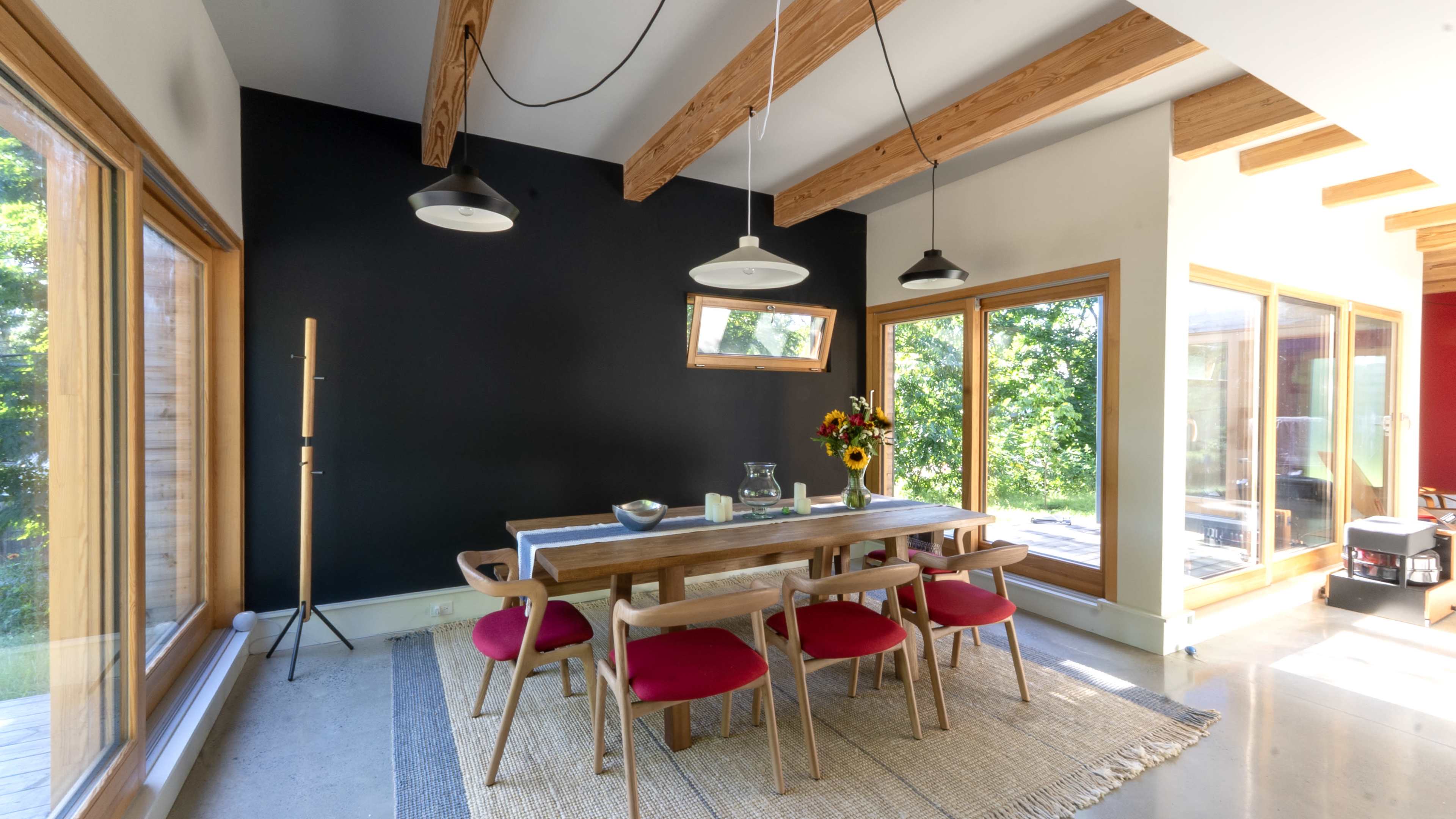 A dining area features a wooden table surrounded by chairs, with large windows allowing natural light to fill the space and a black accent wall adding contrast.