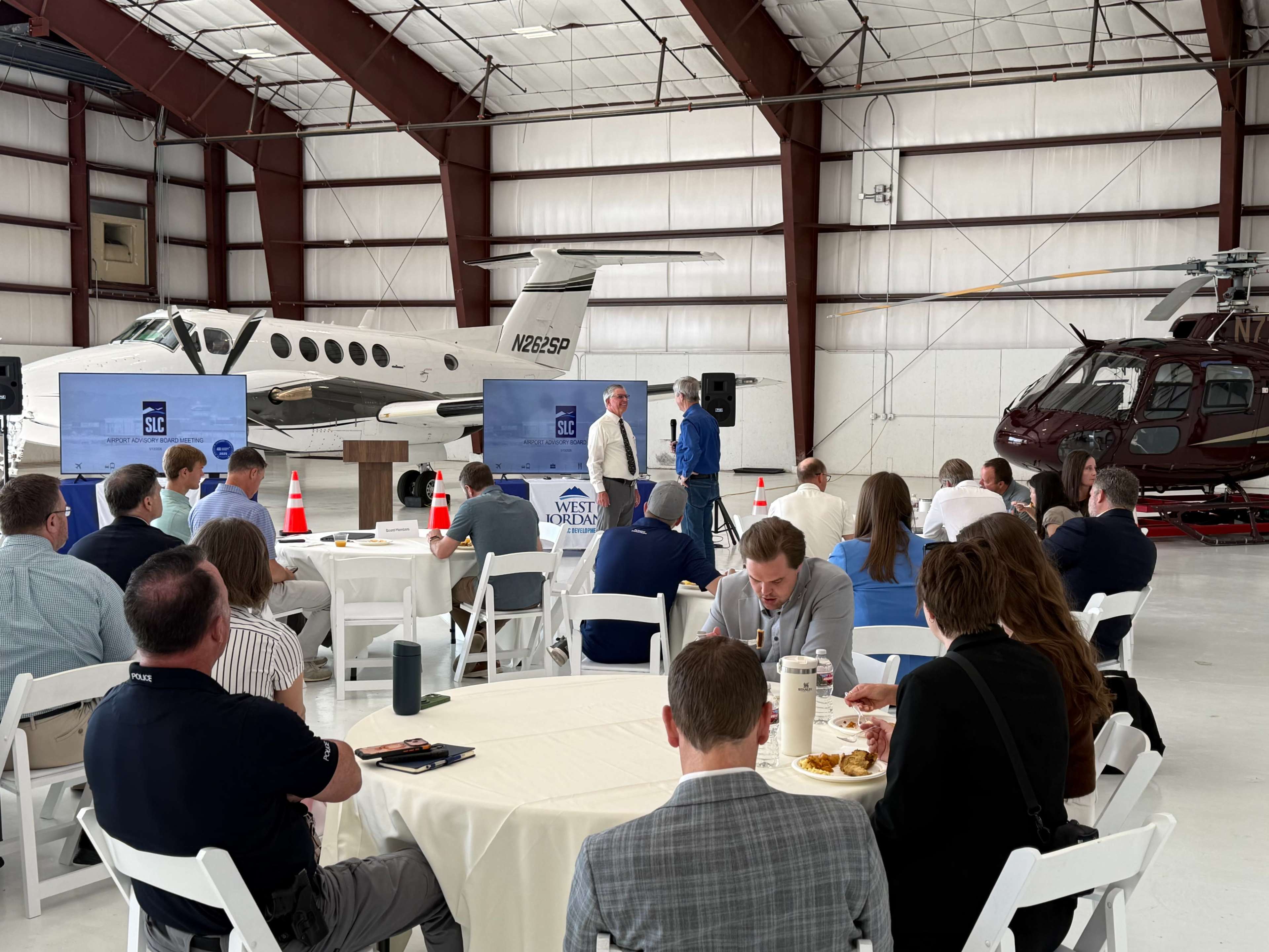 A group of people attend a presentation in a large hangar featuring two airplanes and a helicopter in the background.
